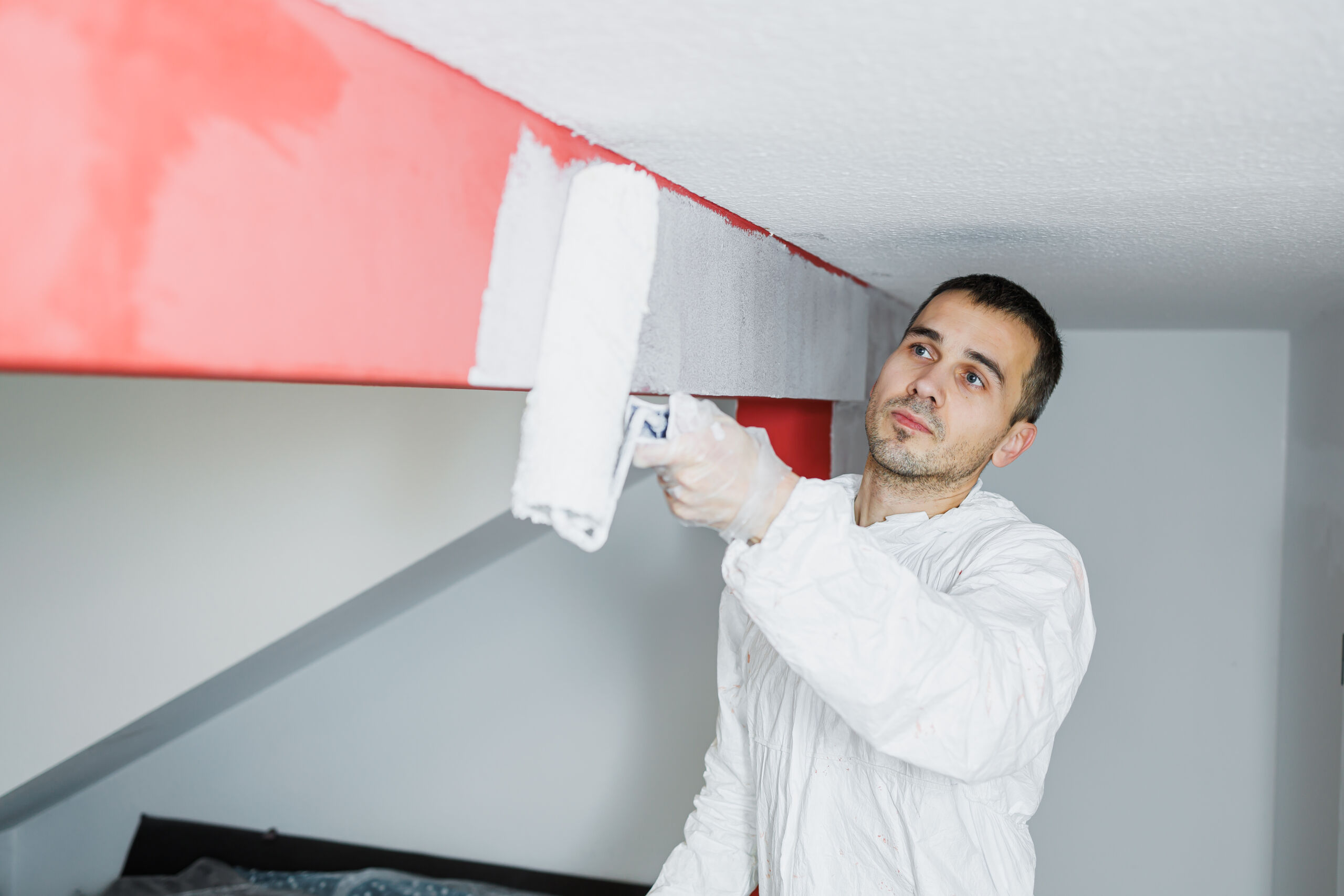 A man wearing a white protective suit paints over a red beam with white paint using a roller in a modern interior space.