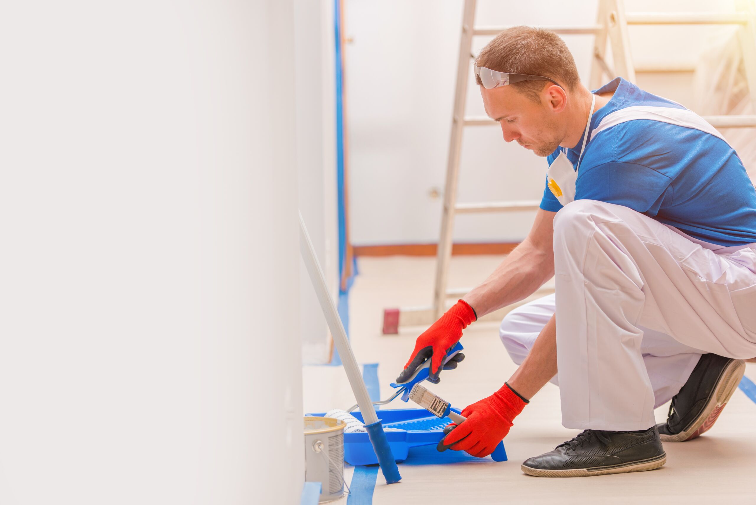 A person wearing gloves and overalls kneels on the floor, dipping a paintbrush into a paint tray; a ladder is visible in the background.