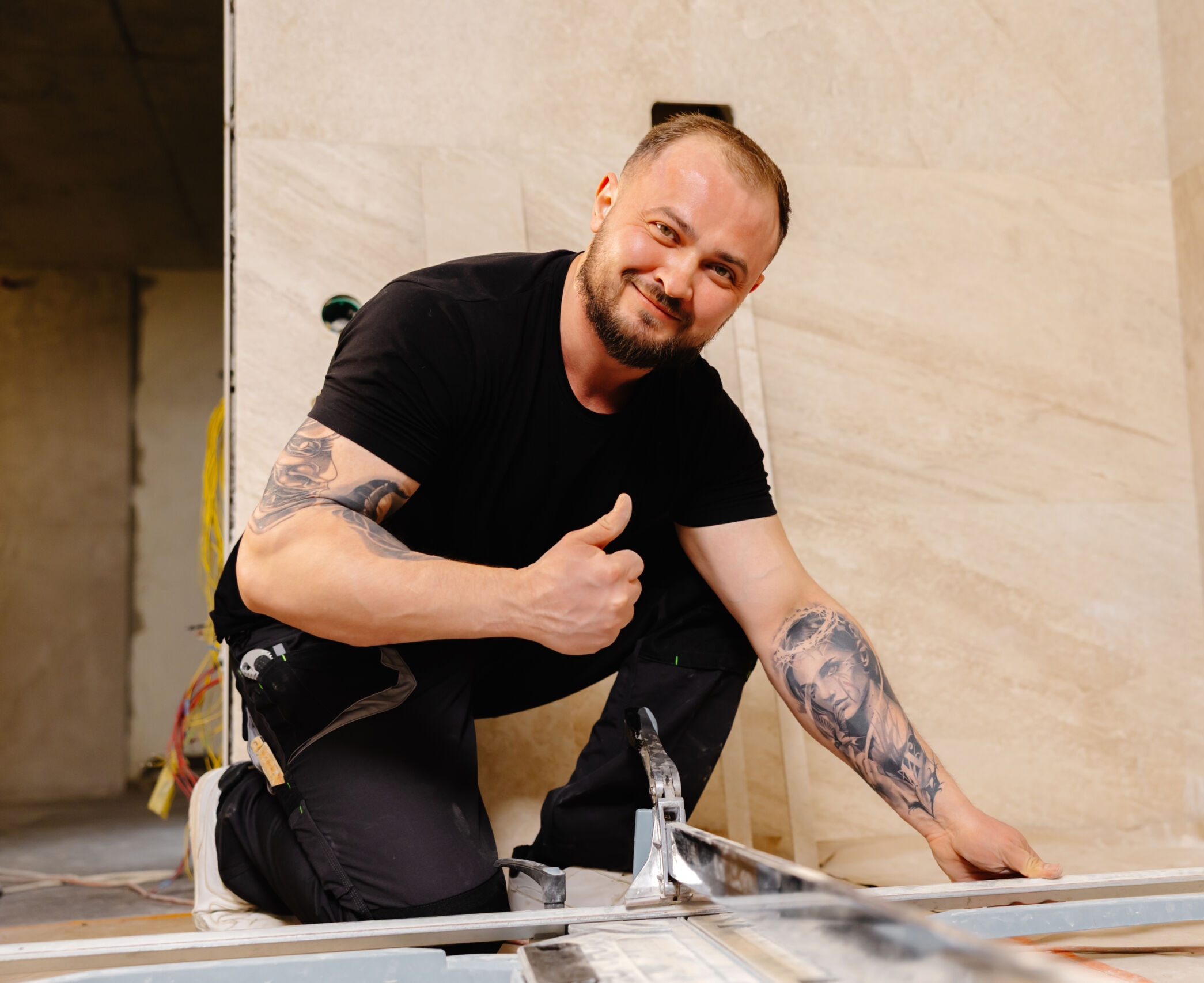 Man kneeling on the floor with construction tools, giving a thumbs up and smiling, in a partially finished room with tiled walls.
