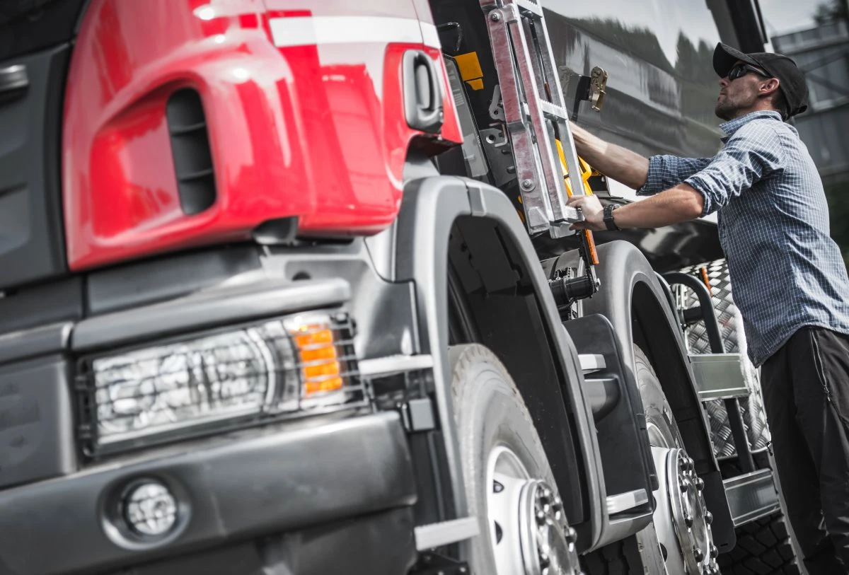 A person in a checked shirt loads or unloads supplies from the side compartment of a large red and black lorry parked outdoors.