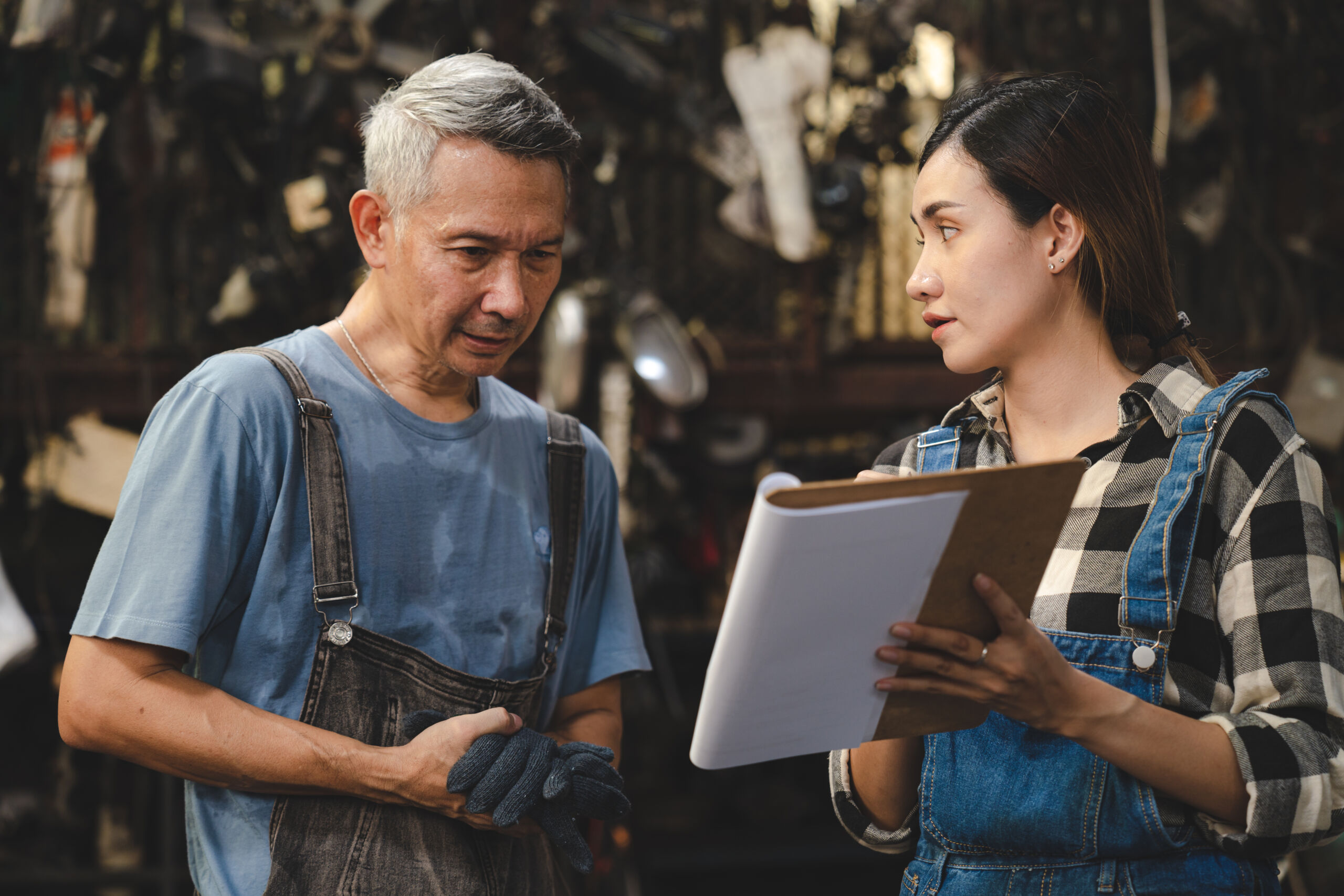 A woman holding a clipboard talks to a man wearing work gloves and overalls. They appear to be in an industrial or workshop setting.
