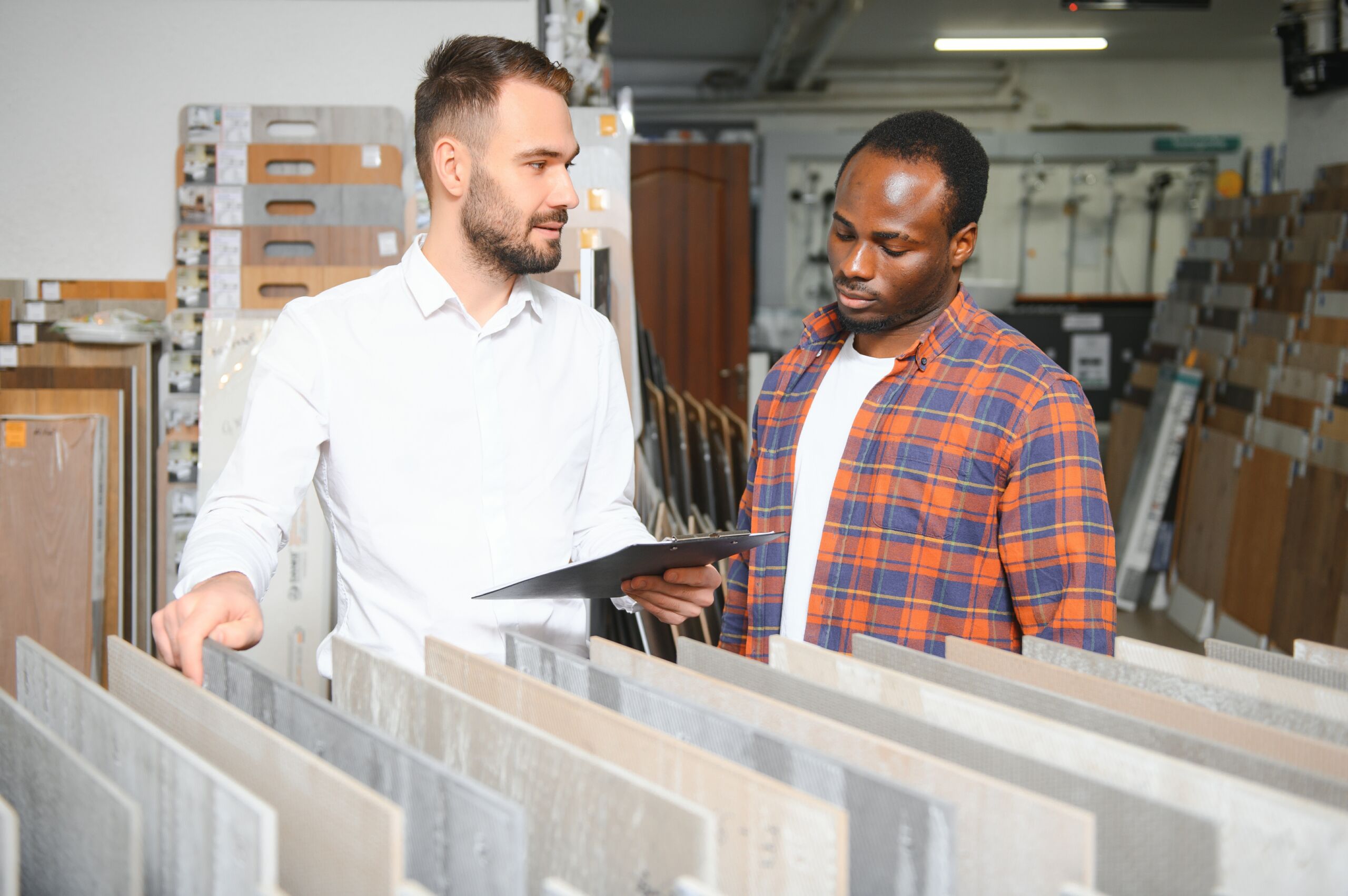 Two men stand in a flooring shop, one holding a clipboard and talking while the other listens, surrounded by sample boards of various flooring materials.