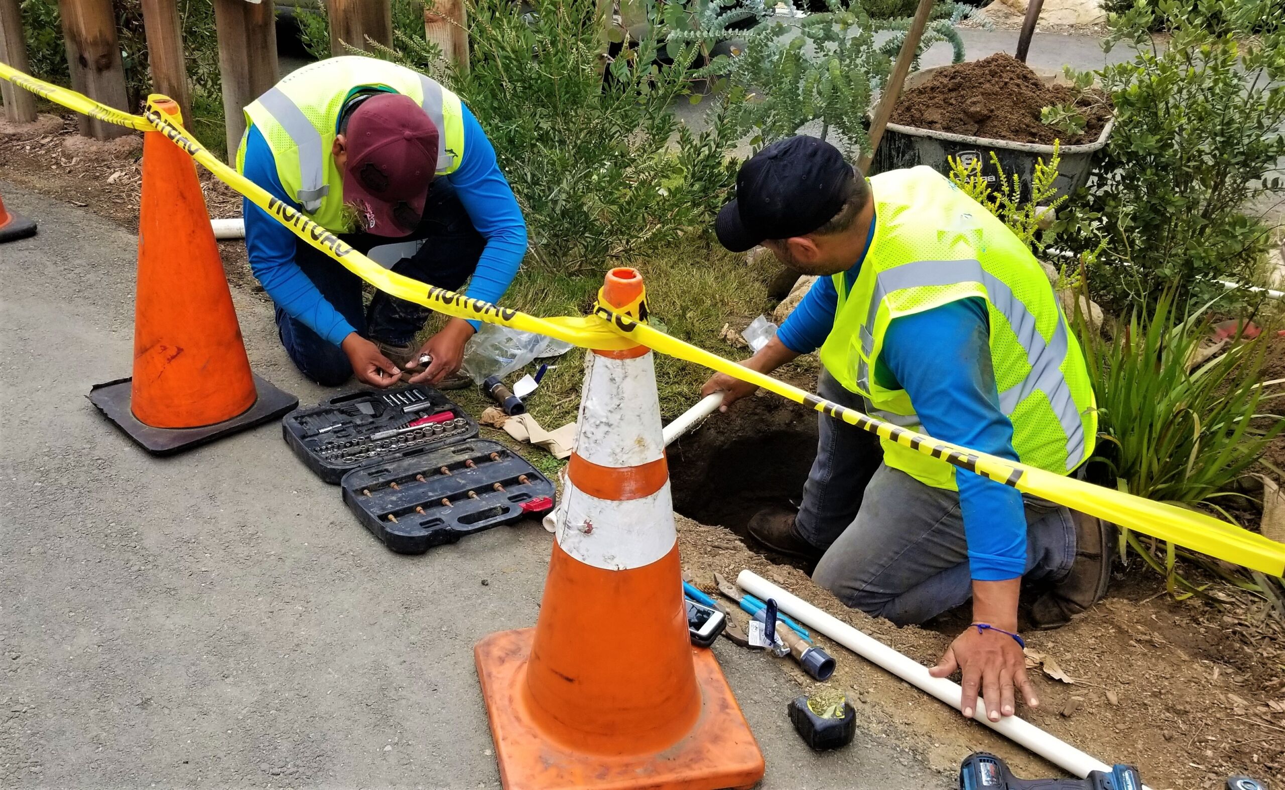 Two workers in safety vests repair pipes beside a road, surrounded by tools, traffic cones, and warning tape.