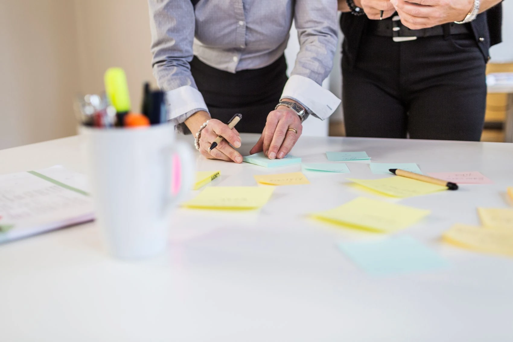 Two people standing at a table covered with colourful sticky notes; one person is writing on a note, and a cup holding pens and highlighters is in the foreground.