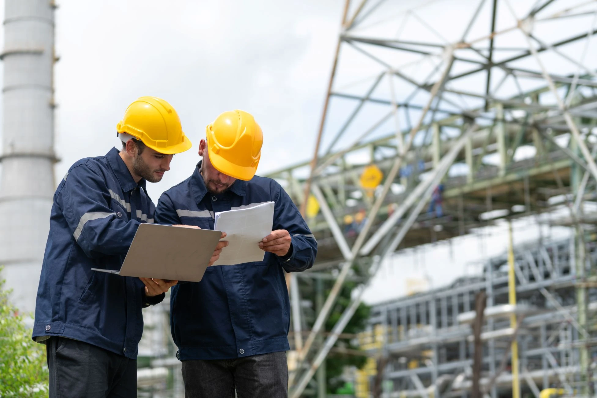 Two workers in hard hats and uniforms review documents and a laptop at an industrial construction site with scaffolding and metal structures in the background.