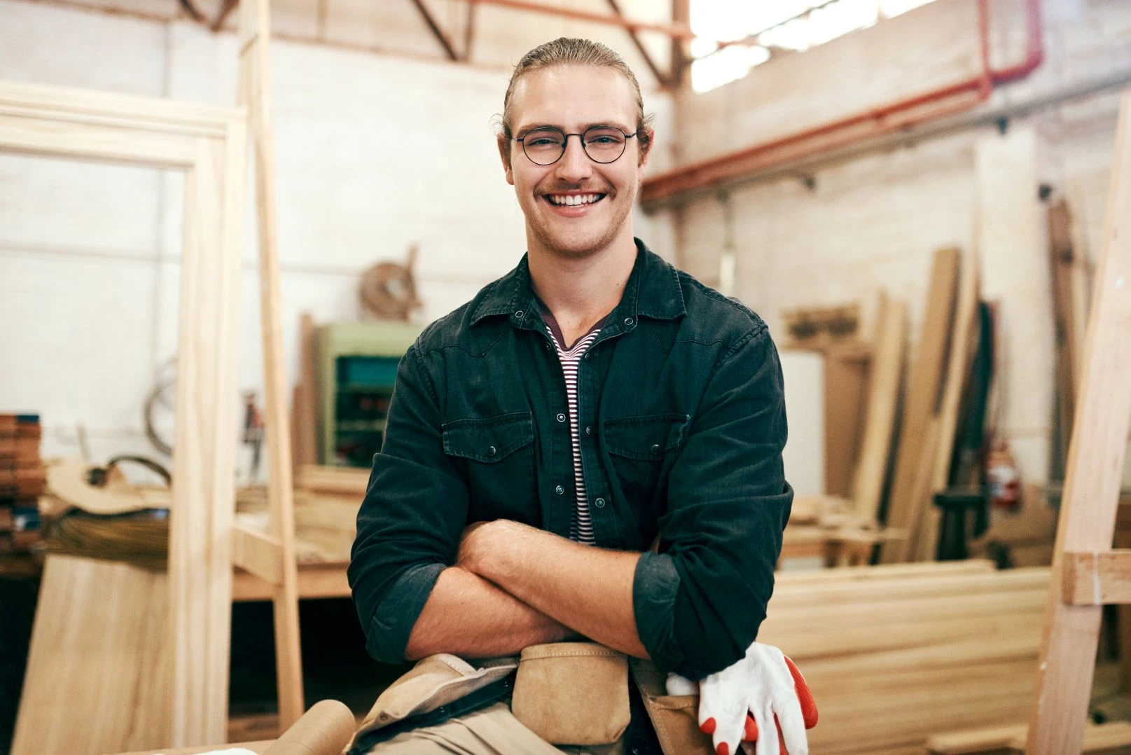 Man with glasses and a moustache, wearing a dark shirt and tool belt, stands with arms crossed in a wood workshop, surrounded by wooden planks and tools.