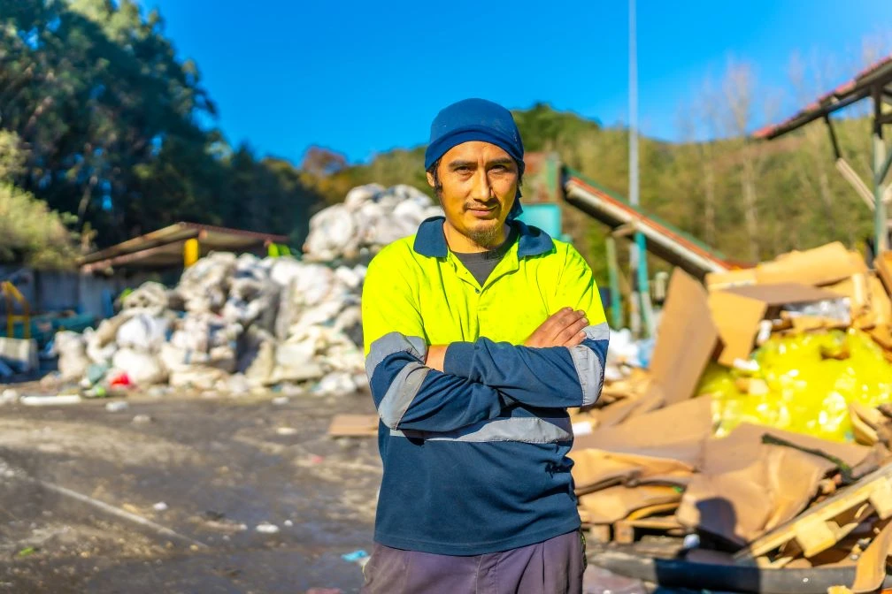 A man in a high-visibility shirt stands with arms crossed at a recycling or waste facility, with piles of cardboard and bags in the background.