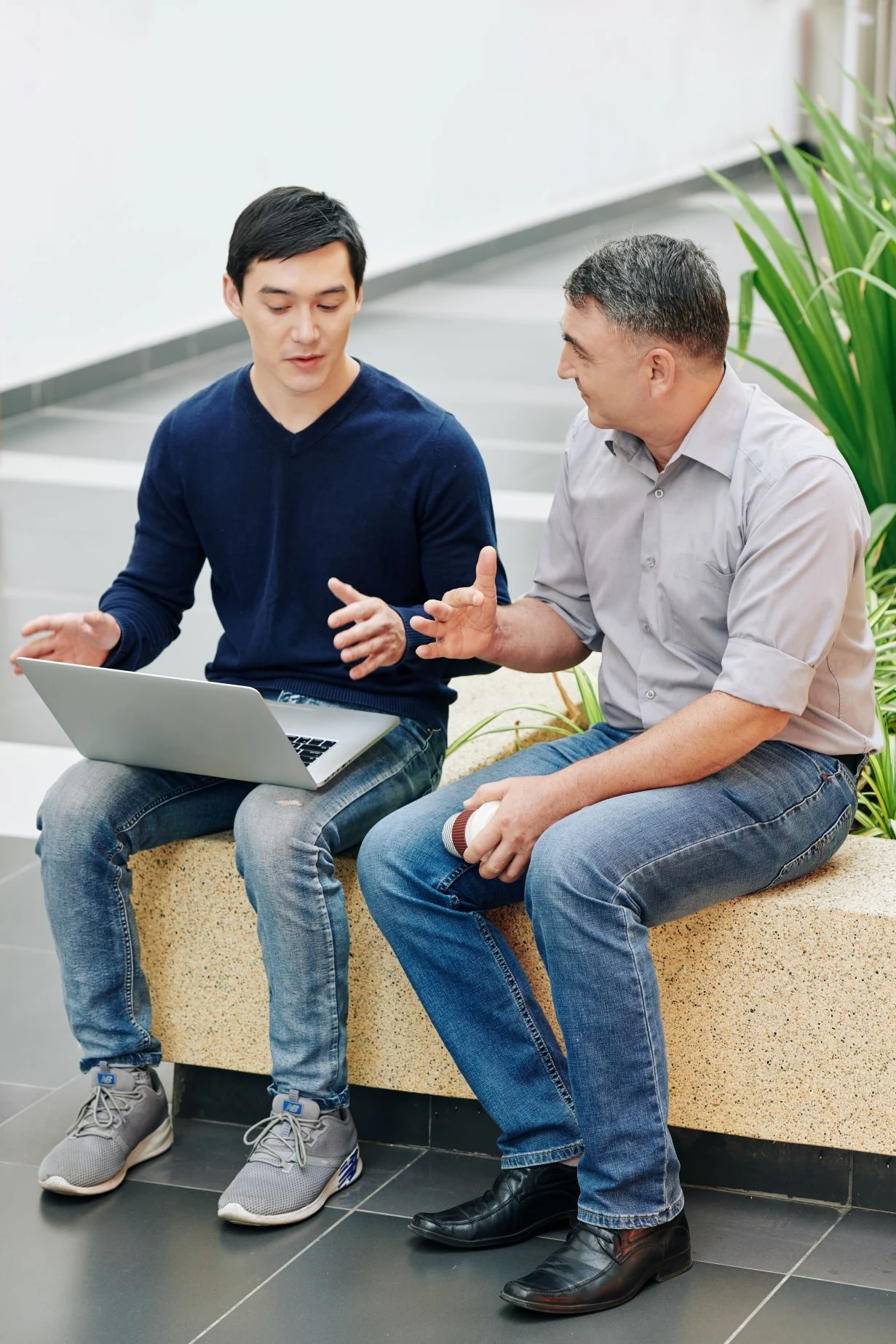 Two men sit on a bench indoors, talking. One has a laptop on his lap, whilst the other holds a white object. Both appear engaged in conversation. Green plants are in the background.