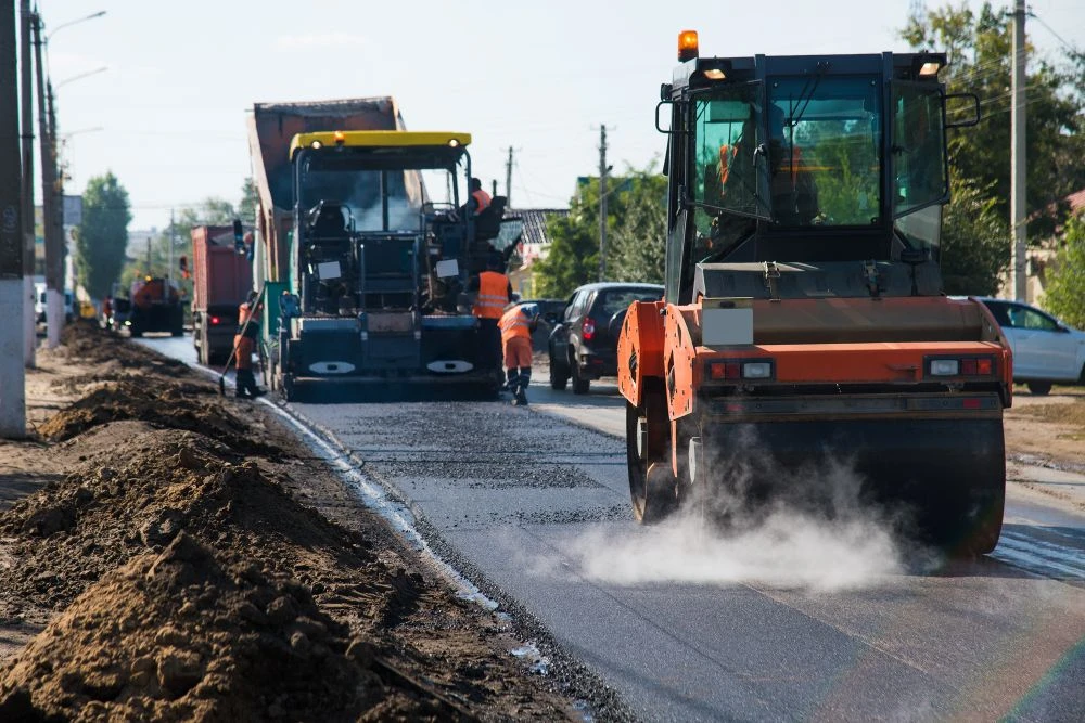 Construction workers and machinery surfacing a road, with a road roller compacting fresh tarmac and vehicles passing nearby.