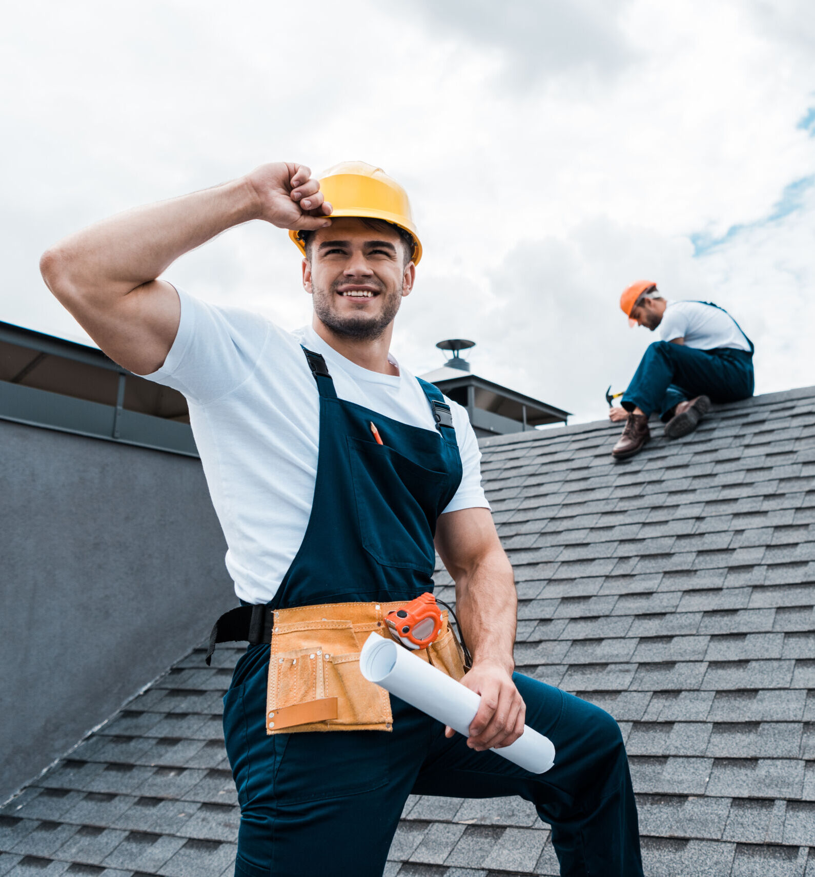 Two construction workers in safety gear are on a tiled roof; one stands holding plans while the other works near the roof's apex.