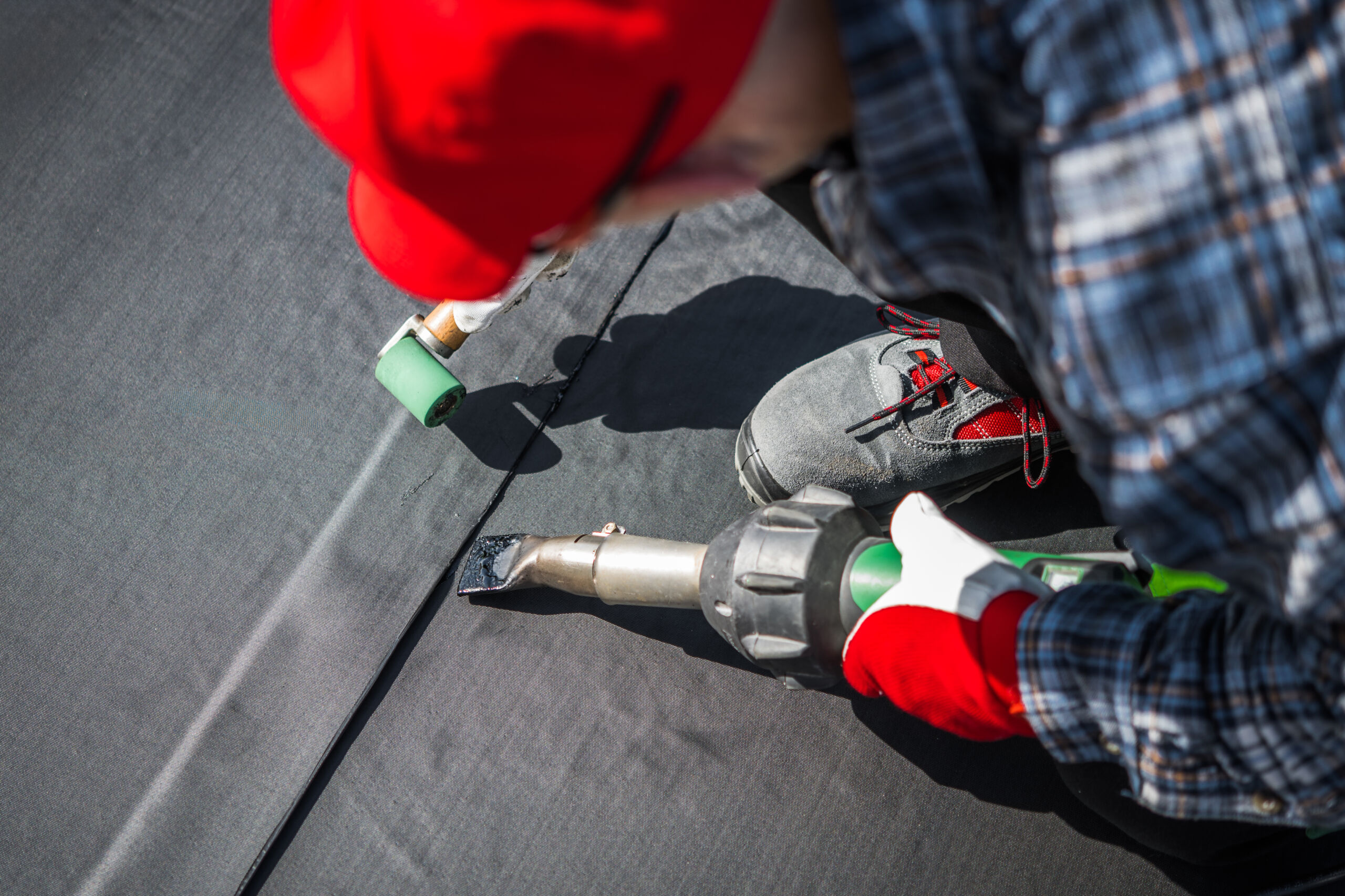 Worker wearing gloves and a red hat uses a heat gun and roller to seal roofing membrane on a flat roof.