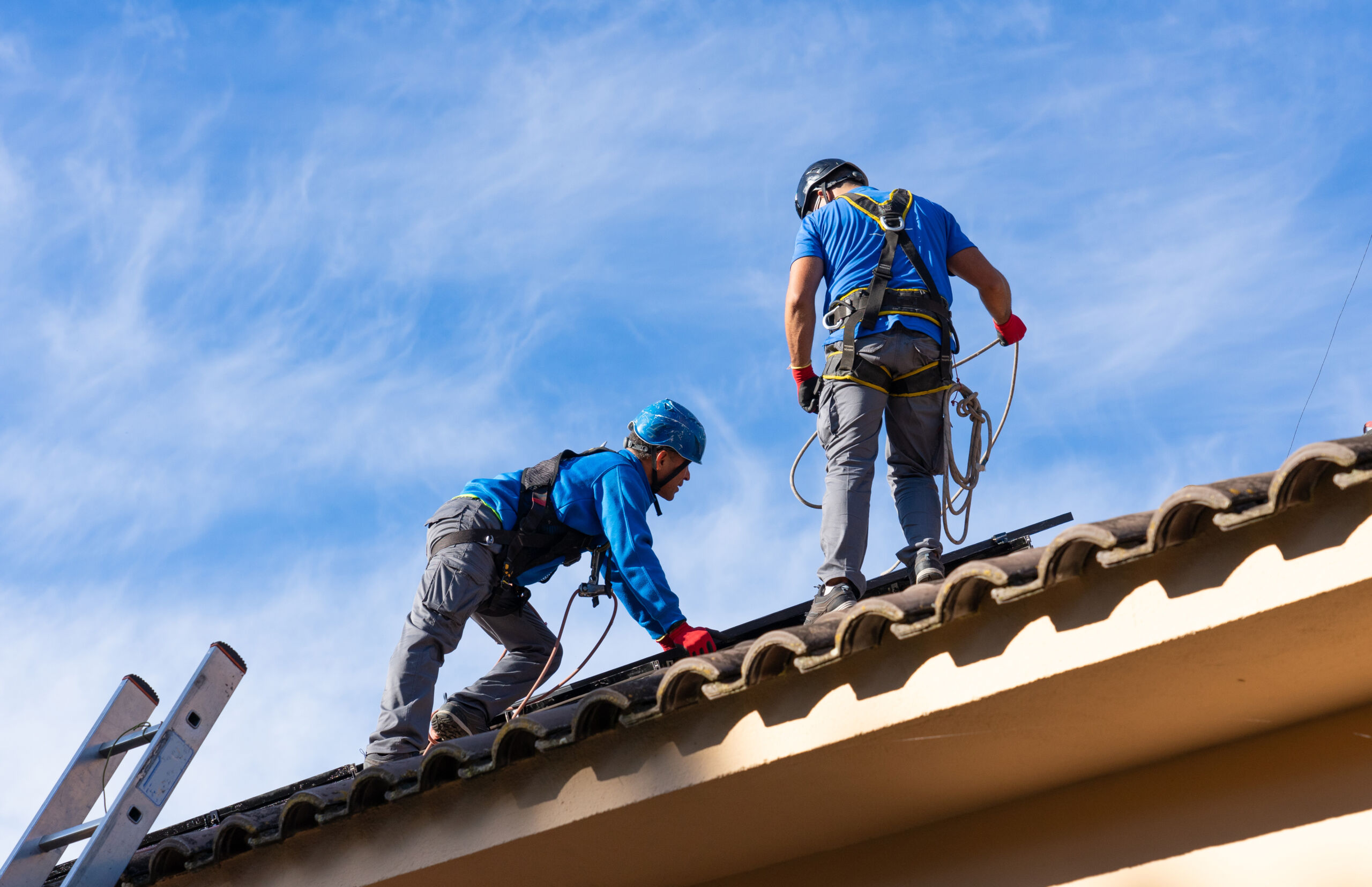 Two workers wearing safety gear are installing or inspecting solar panels on a tiled roof under a blue sky.