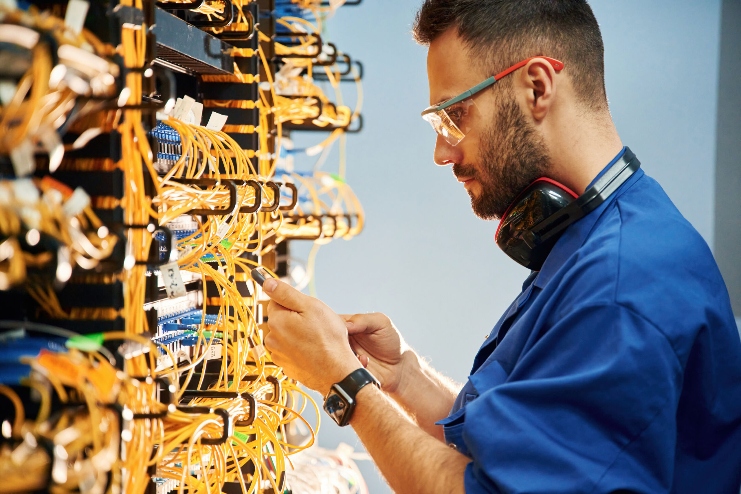A technician in safety spectacles and a blue shirt works on a server rack filled with cables, inspecting connections.