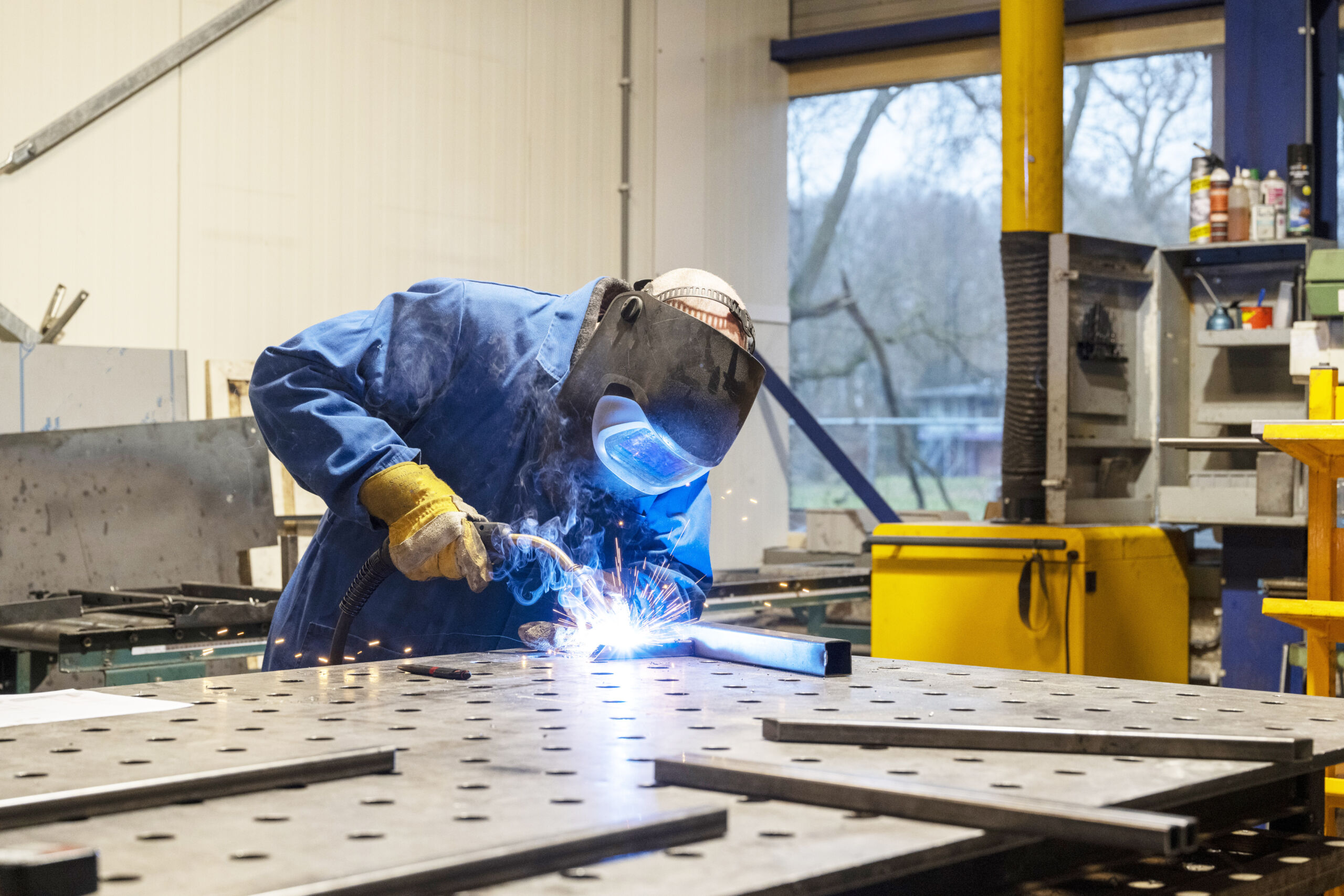 Person in protective gear welding metal on a workbench in an industrial workshop, with sparks visible from the welding process.