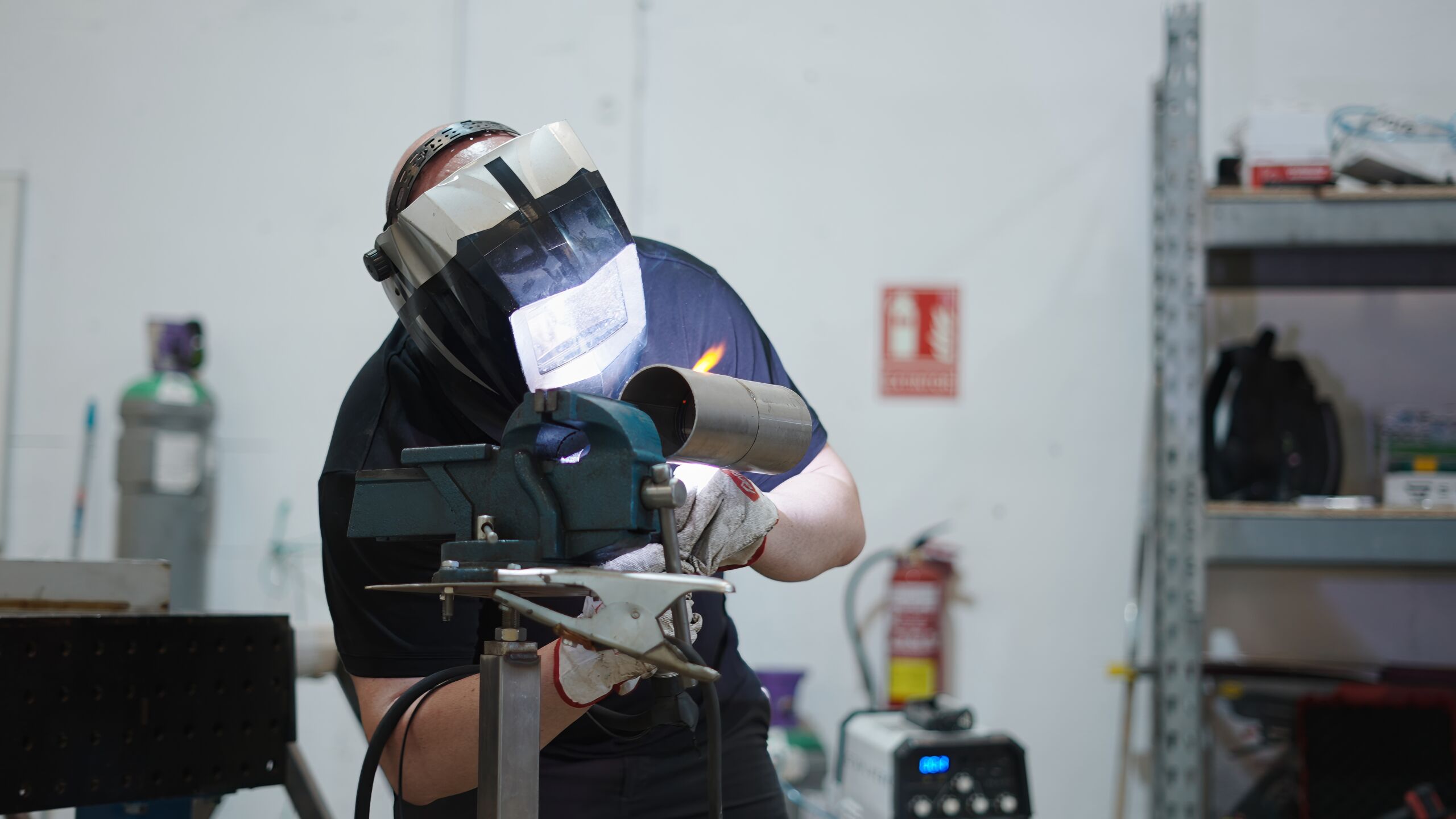 Person wearing a welding helmet and gloves, welding a metal pipe clamped in a vice inside a workshop with various tools and equipment in the background.