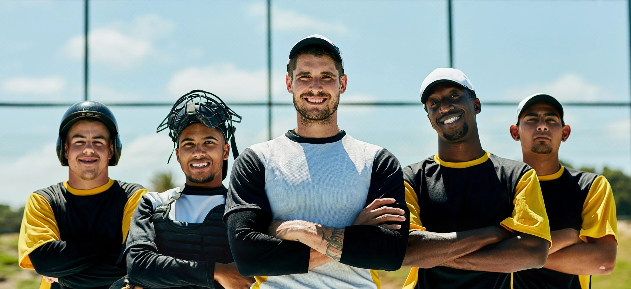 Five adult male baseball players in black and yellow kits stand outdoors in a line, smiling, with arms folded, under a sunny sky.