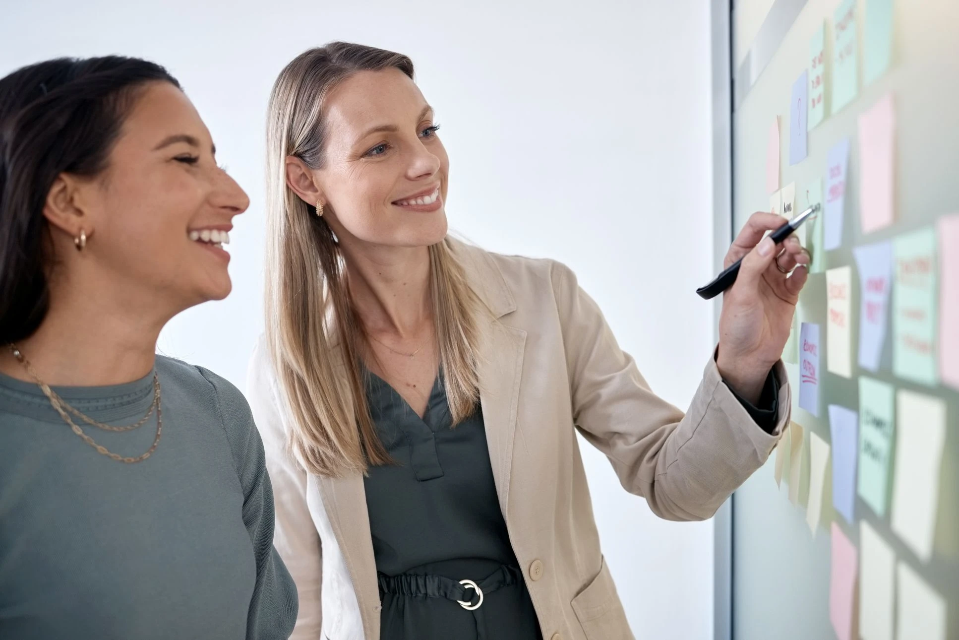Two women stand next to a glass wall covered with sticky notes. One woman points at a note with a marker, and both are smiling.