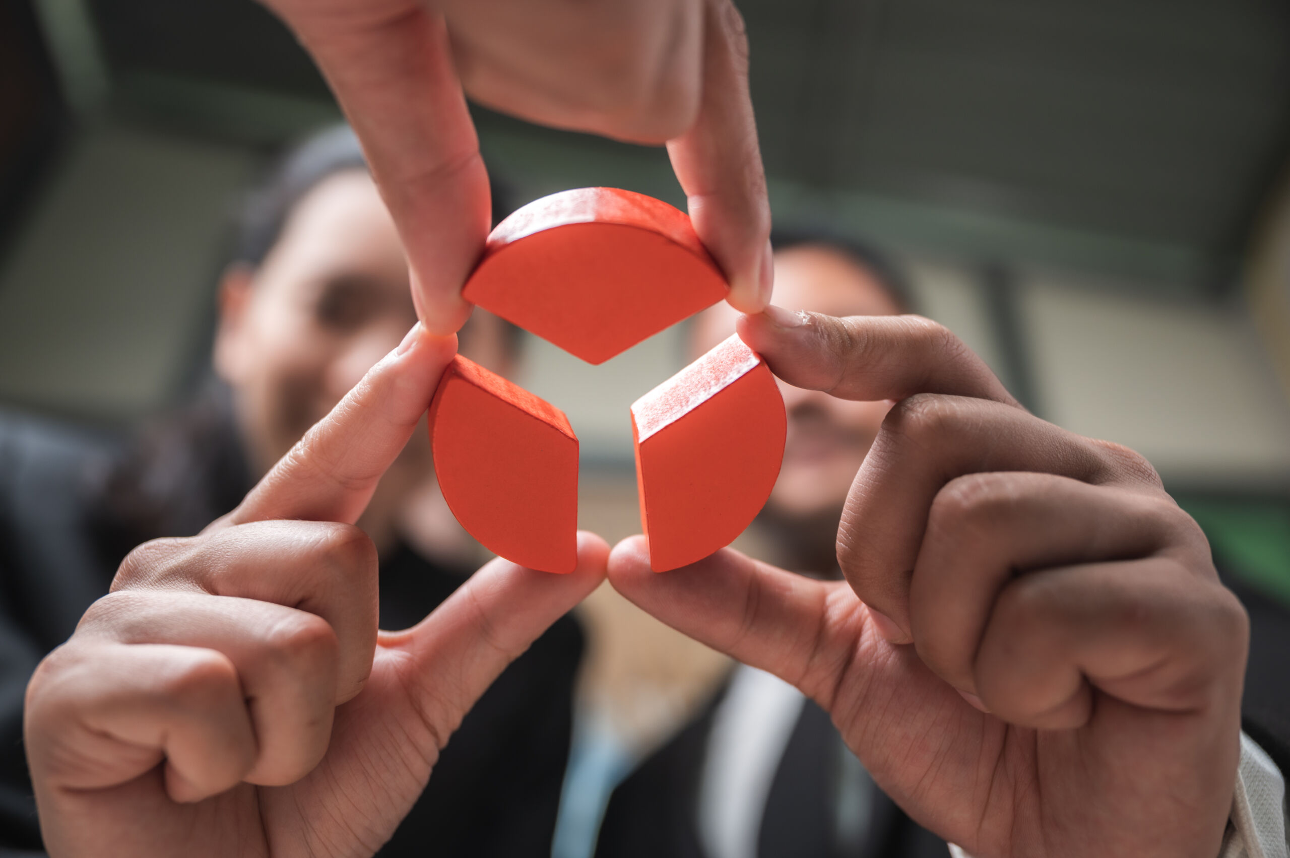 Four hands hold three red jigsaw pieces forming a circle, with two blurred people in the background.