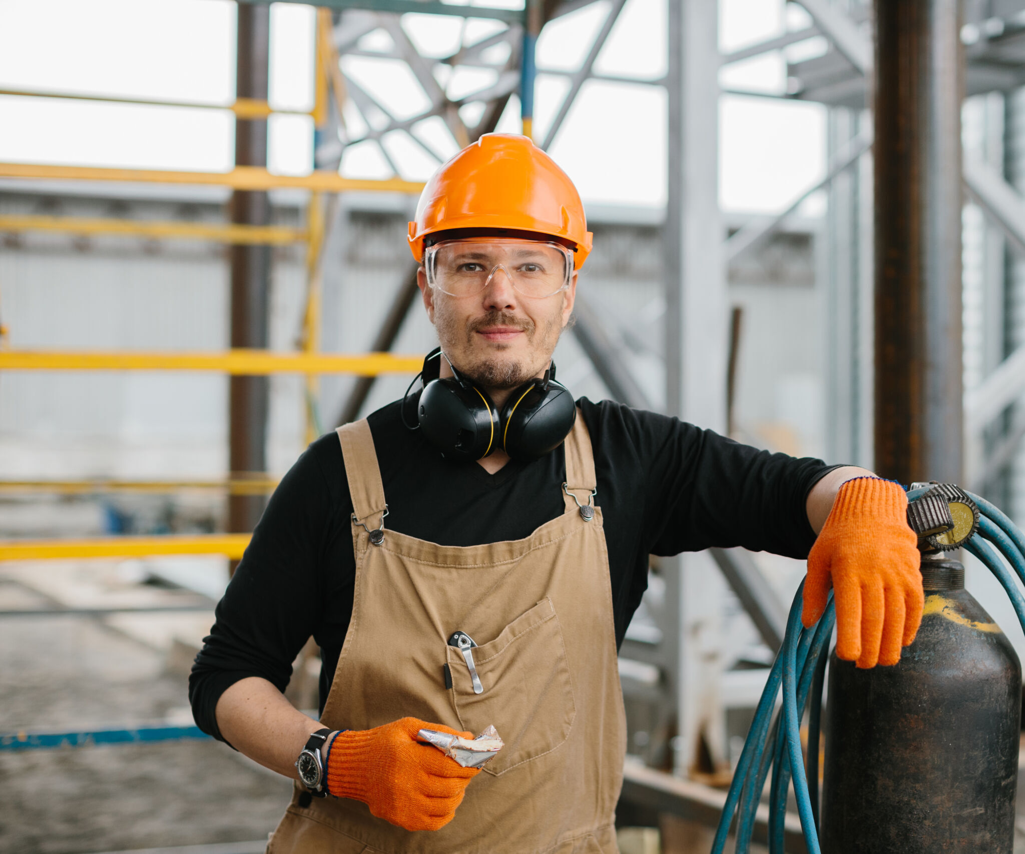 A construction worker in safety gear, including an orange helmet and gloves, stands outdoors holding a tool and leaning on equipment with blue hoses. Steel framework and scaffolding are visible behind.