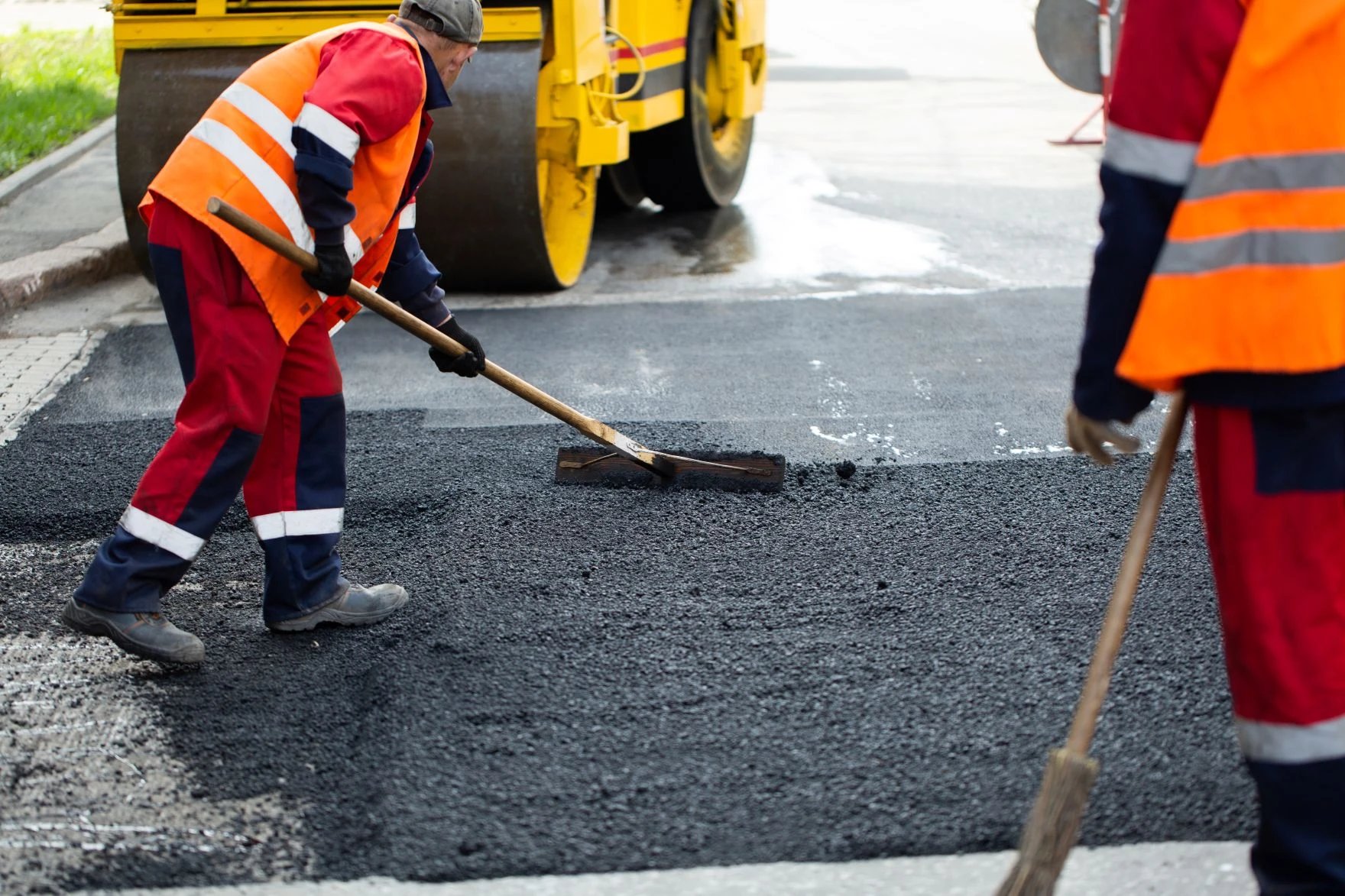 Two workers in safety gear spread asphalt on a road using tools, with a yellow road roller visible in the background.