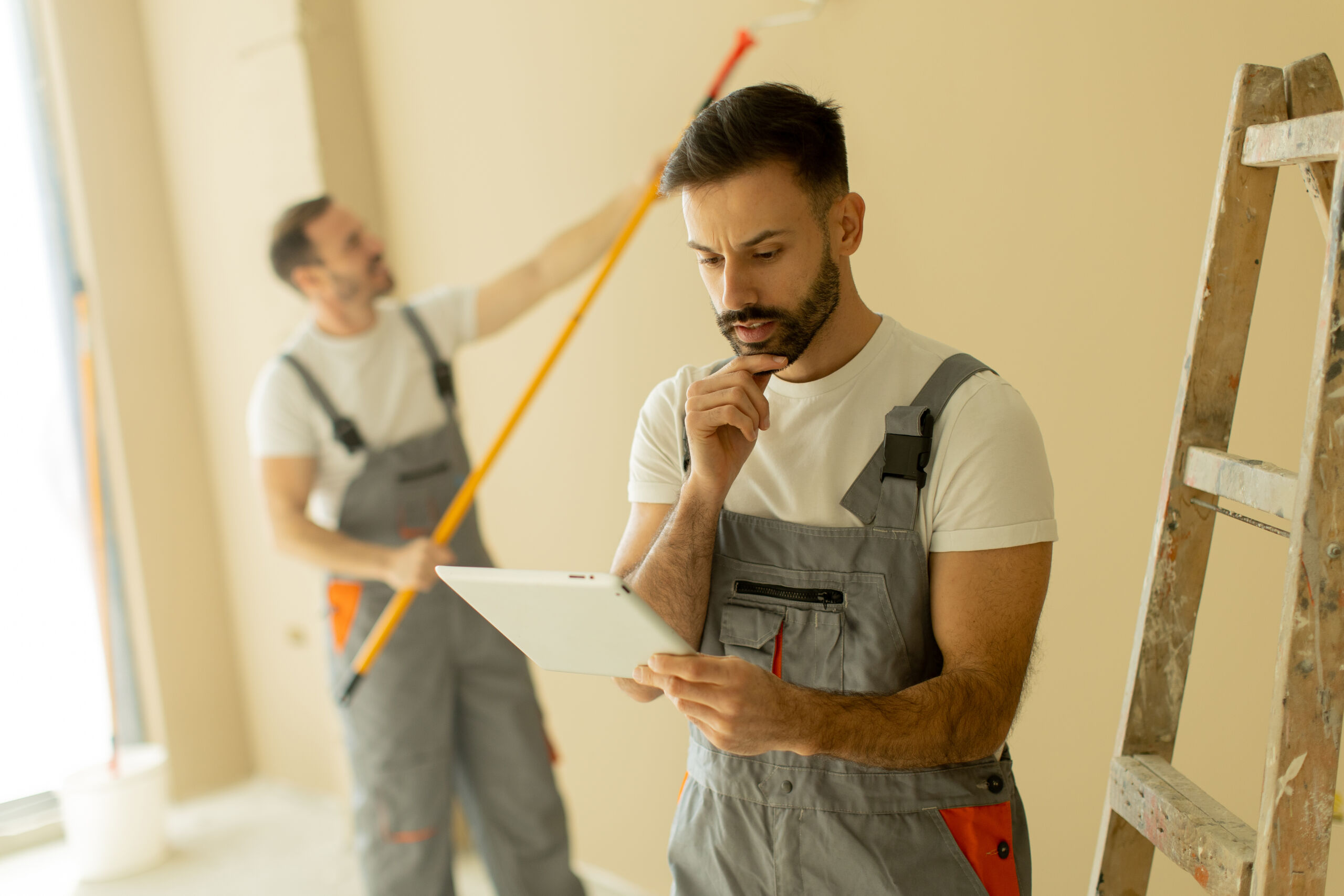 Two men in work overalls; one paints a beige wall with a roller, whilst the other stands near a ladder, looking thoughtfully at a tablet.