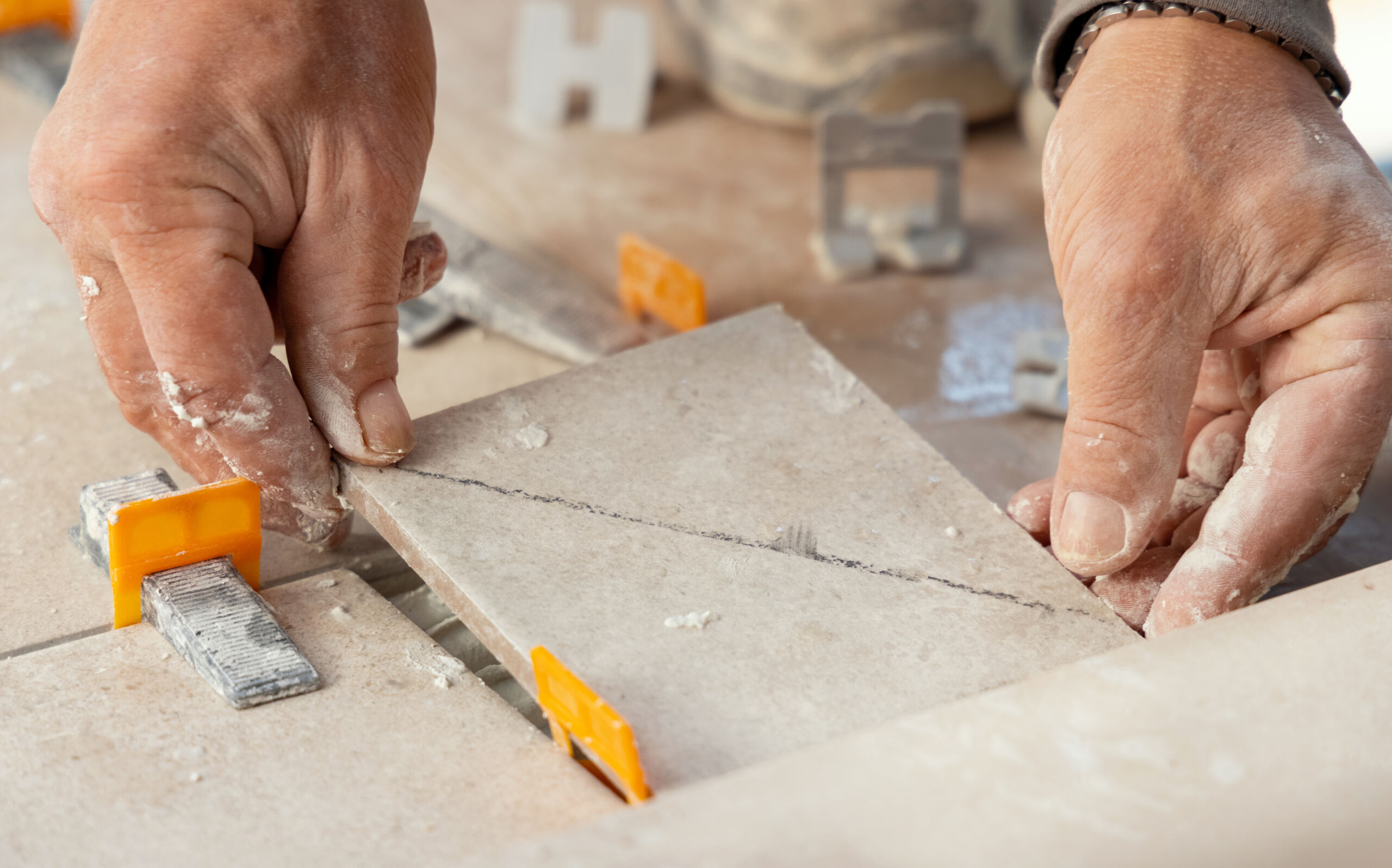 Close-up of hands placing a ceramic tile with a diagonal cut onto a floor, using orange tile spacers and levelling tools.