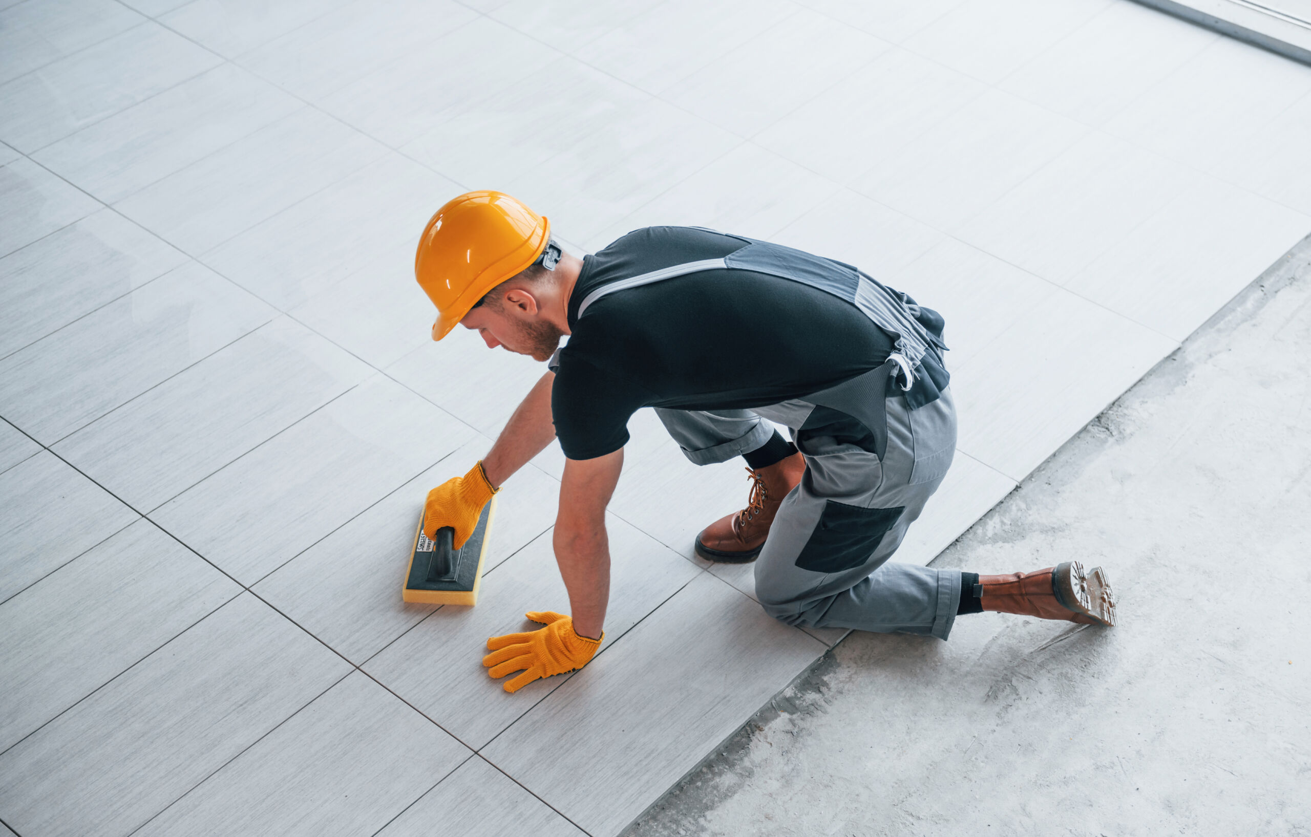 A worker in a hard hat and gloves kneels on the floor, using a tool to clean or finish newly installed tiles.
