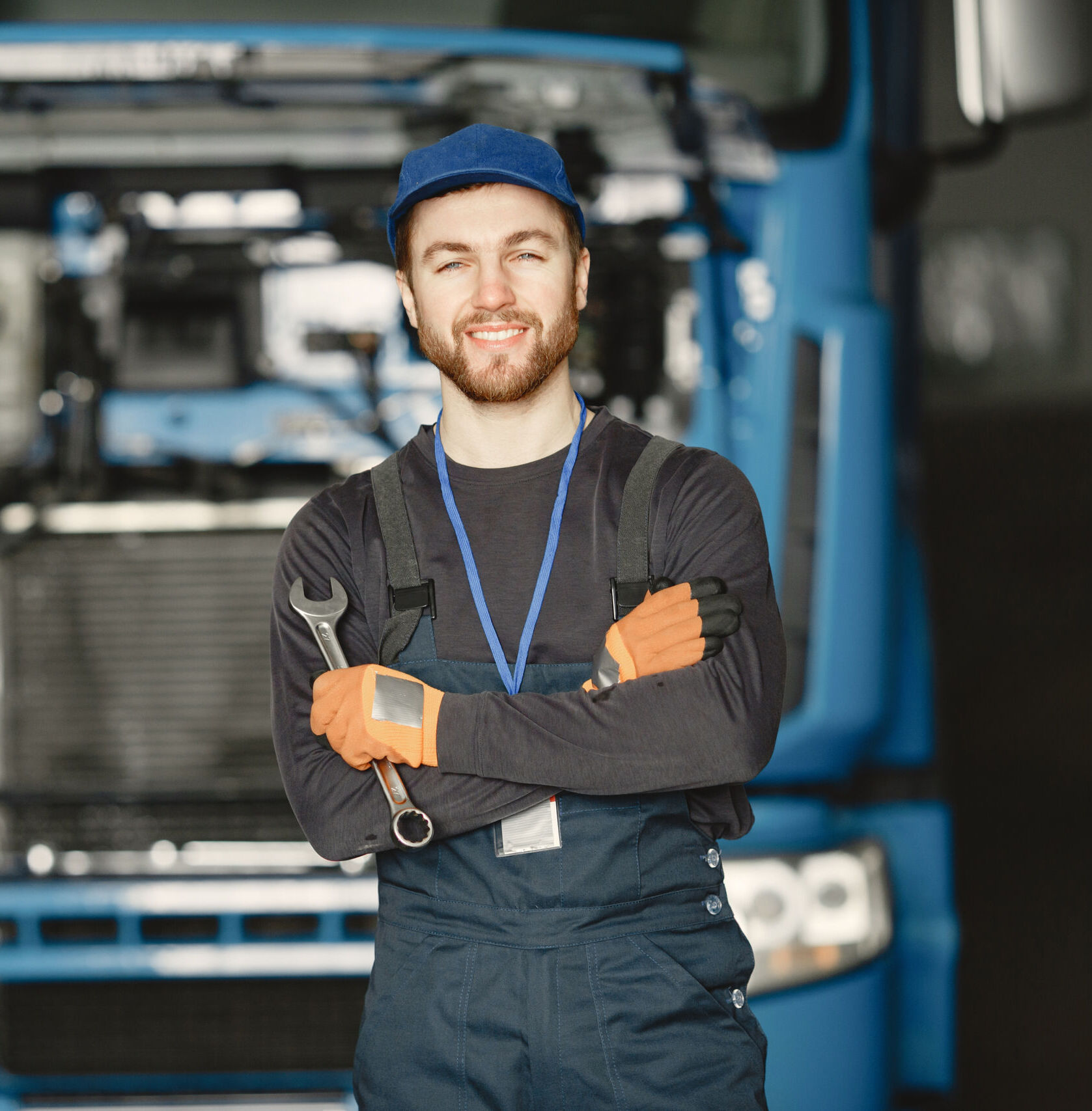 Mechanic in uniform stands with arms folded holding a spanner in front of a blue lorry with its bonnet open, smiling at the camera.