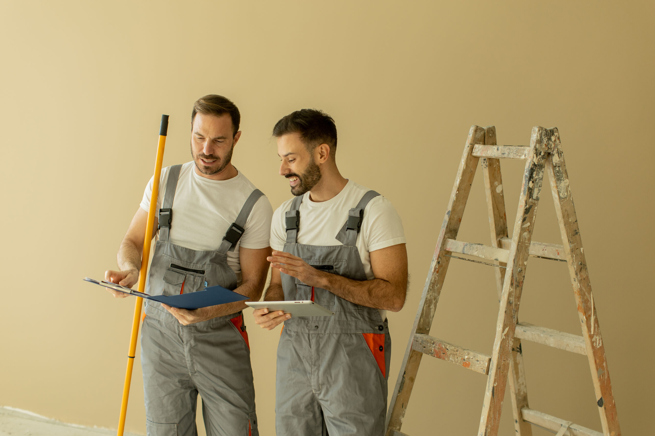 Two men in work overalls review documents next to a yellow post and a wooden ladder against a beige wall.
