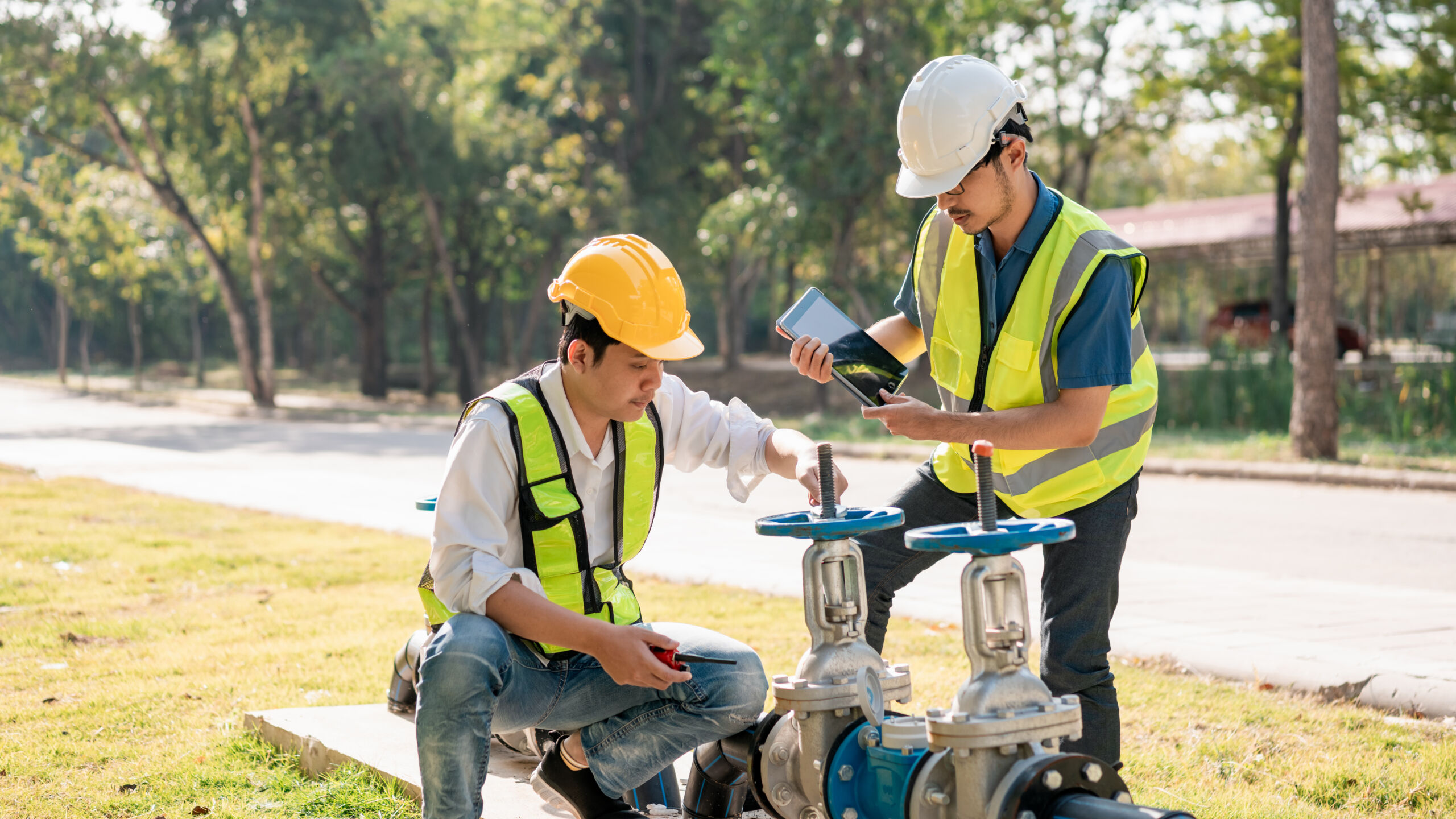 Two workers in safety gear inspect and adjust outdoor valves. One worker is kneeling with a tool, while the other stands, holding a tablet. Trees and a road are visible in the background.