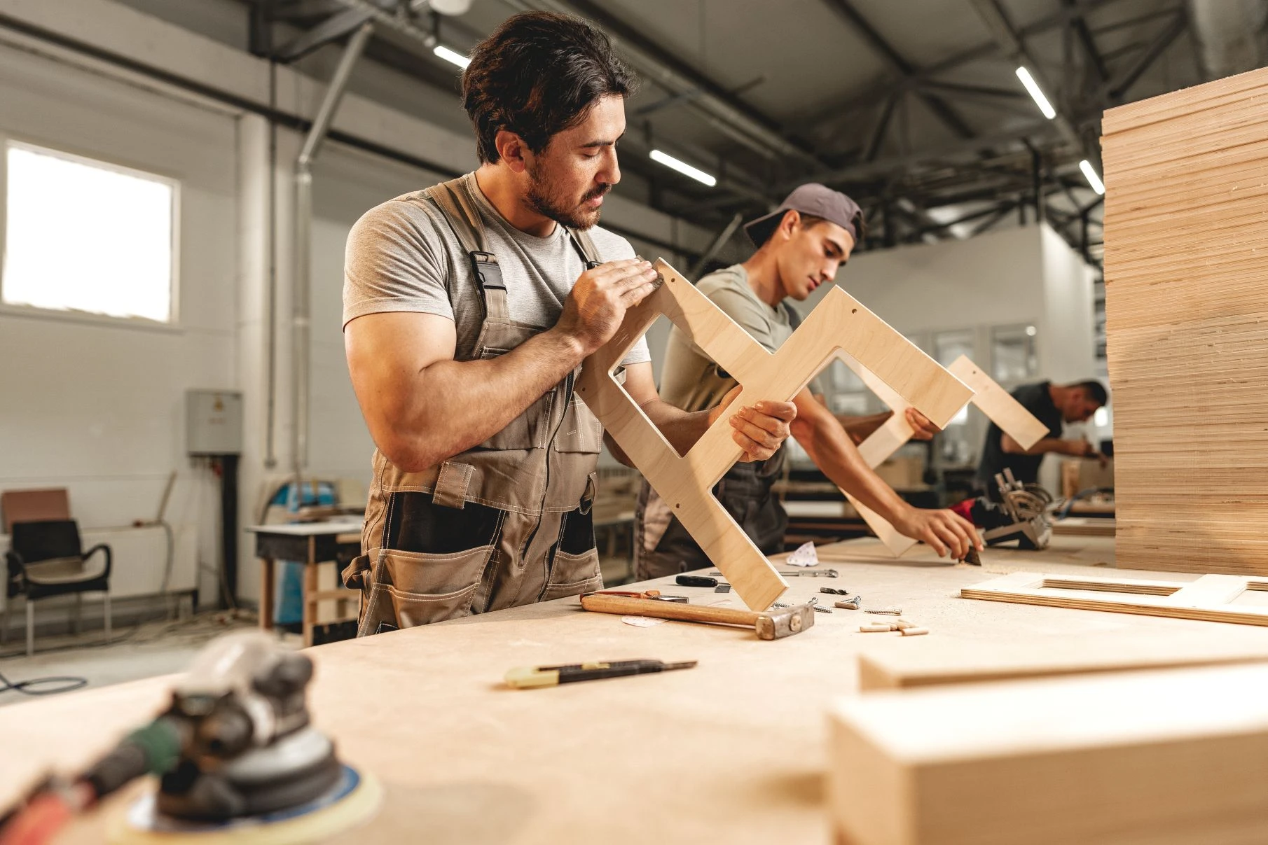 Two men in work overalls assemble wooden furniture pieces on a workbench in a well-lit carpentry workshop. Various tools and wood materials are visible on the table.