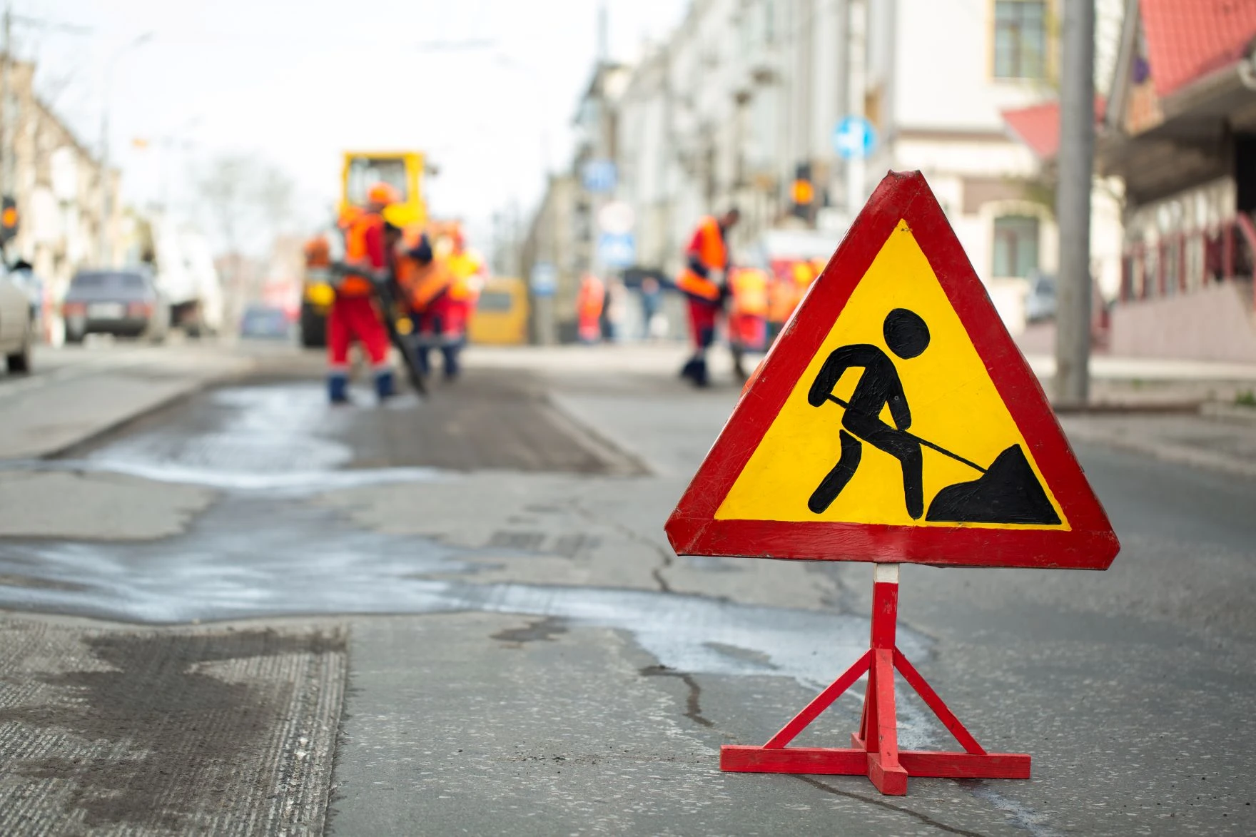 Triangular roadworks sign in foreground with construction workers working on a city street in the background.