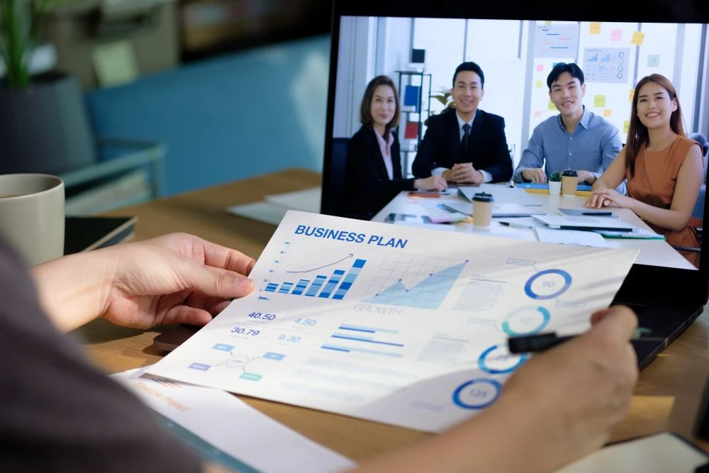 Person reviews a business plan chart whilst attending a video conference with four colleagues displayed on a laptop screen.