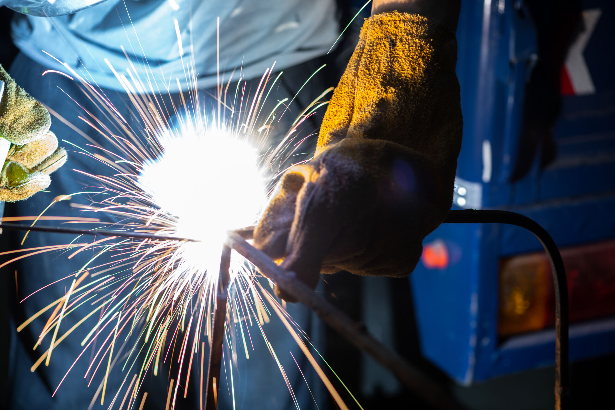 A person wearing protective gloves welds metal, producing bright sparks and intense light.