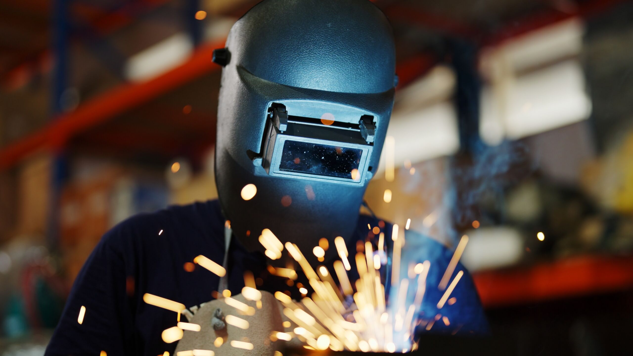 Person wearing a welding helmet is welding metal, with bright sparks flying in the foreground.