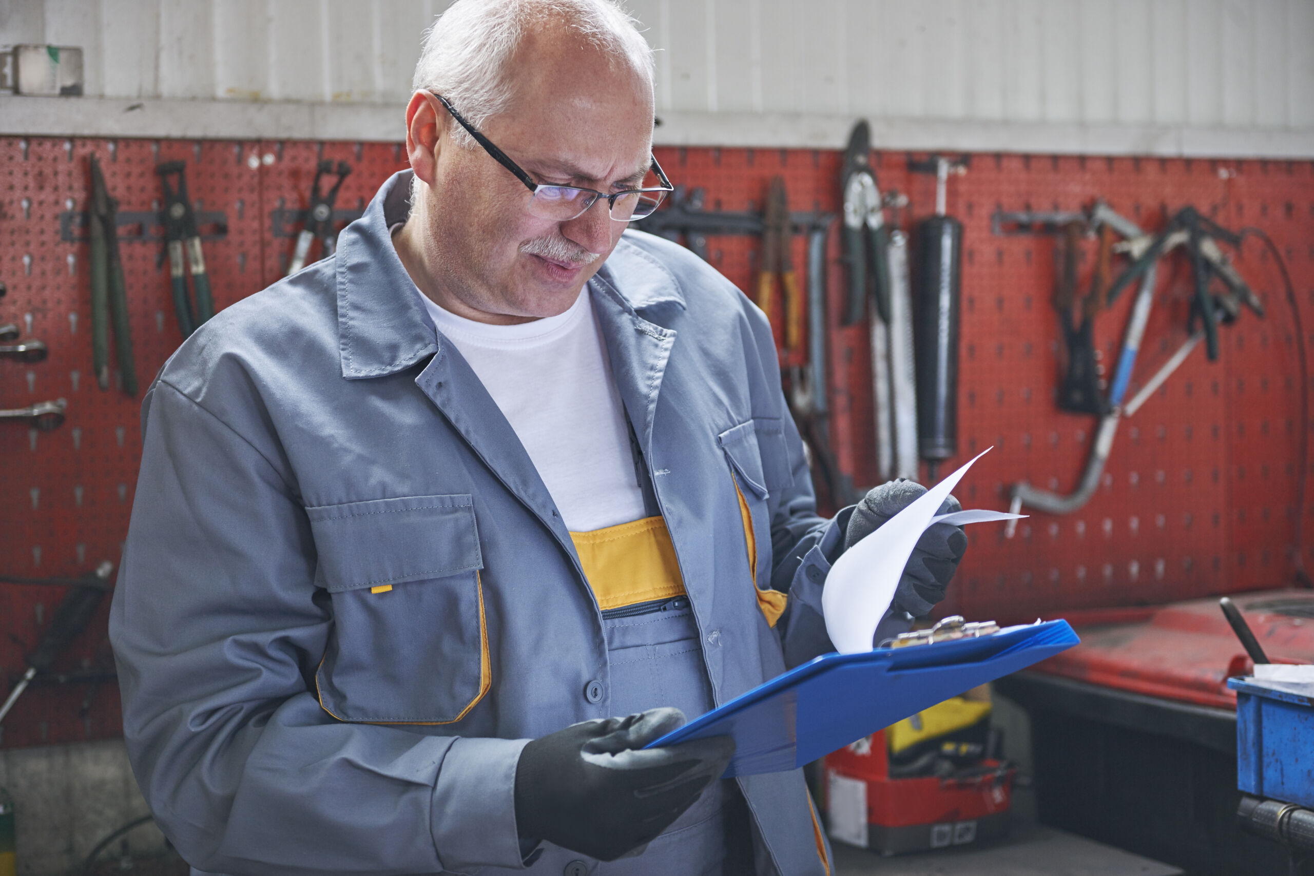 Older man in work overalls and gloves examines papers on a clipboard in a workshop with various tools hanging on a red pegboard wall.