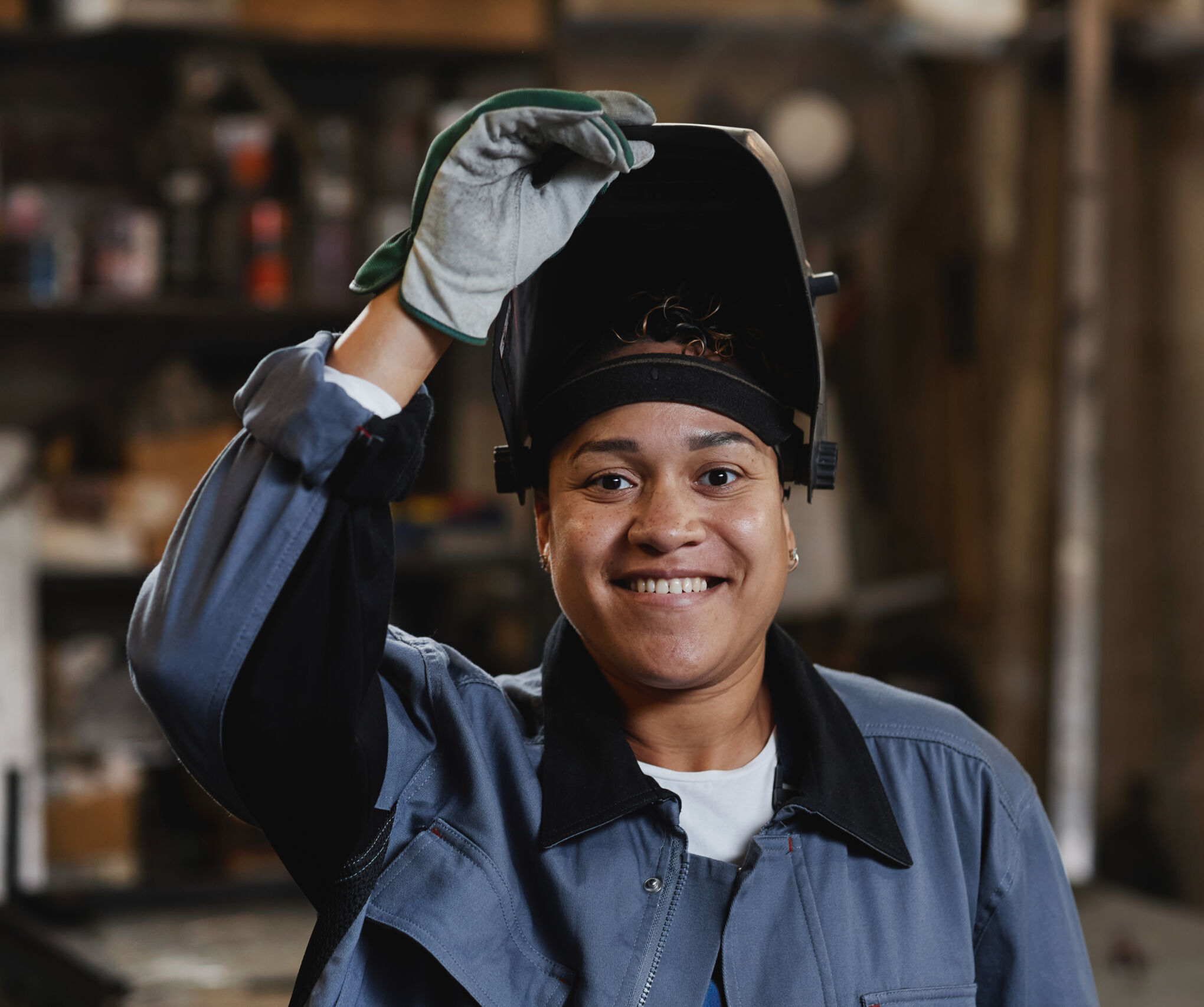 Person in workwear and gloves lifts welding helmet, smiling, in an industrial workshop setting.