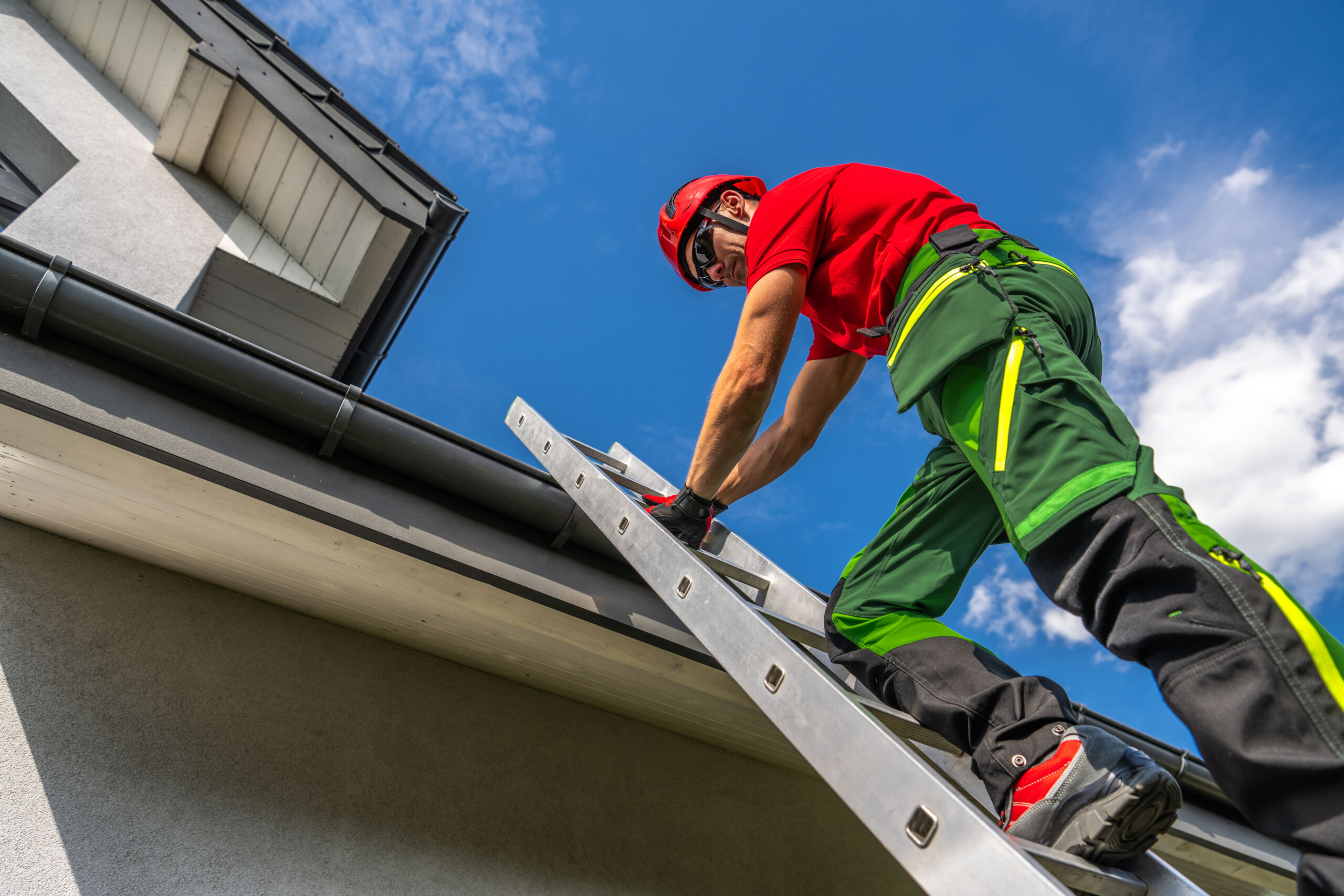 Worker in safety gear stands on a ladder and repairs or inspects the gutters on a house exterior under a clear blue sky.