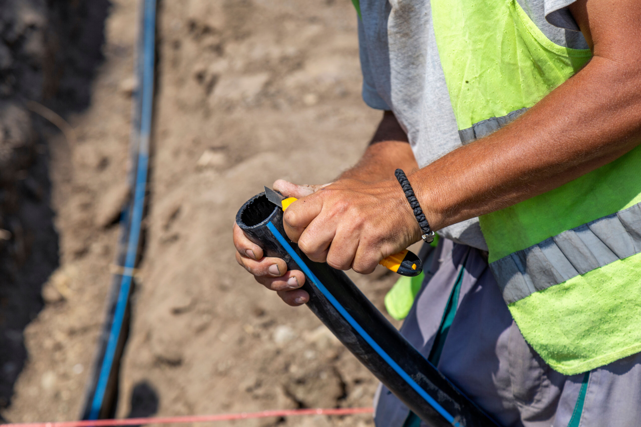 A worker wearing a safety vest uses a utility knife to cut a black plastic pipe at a construction site with visible soil and trench.