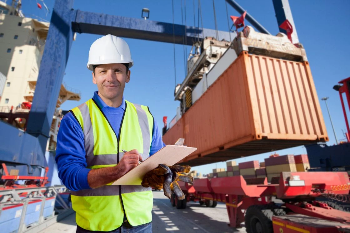 A construction worker in a hard hat and safety vest holds a clipboard, standing in front of a shipping container being lifted by a crane at a busy shipping yard.