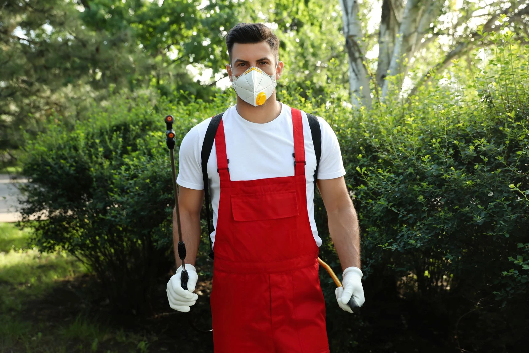 Man in red overalls and white shirt wearing a protective mask and gloves, holding gardening tools, standing outdoors near green bushes.