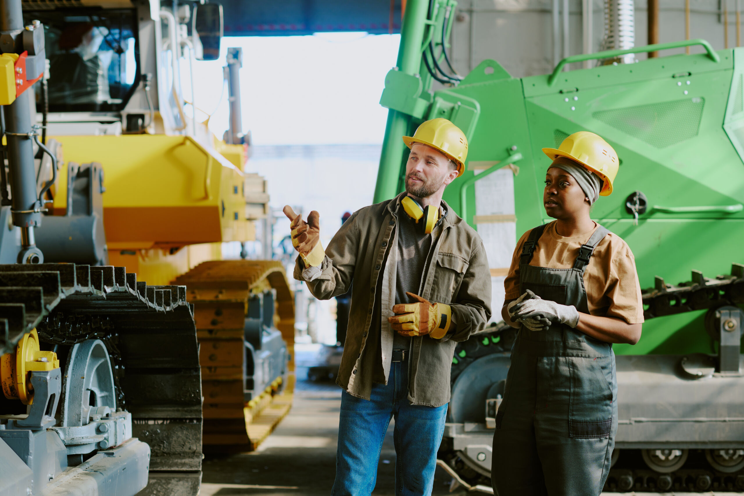 Two construction workers in safety gear stand indoors, discussing near large machinery, including a green vehicle and a yellow digger.