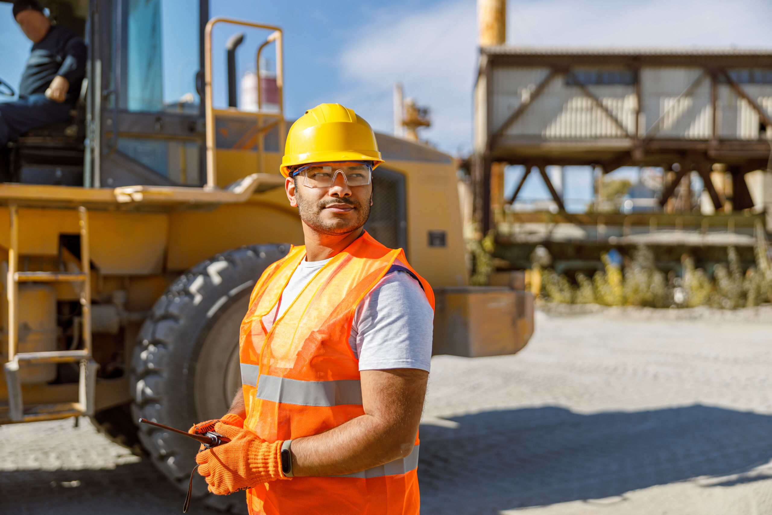 Man wearing a yellow hard hat, orange safety vest, and gloves stands outdoors at a construction site, holding a tablet, with heavy machinery and industrial structures in the background.