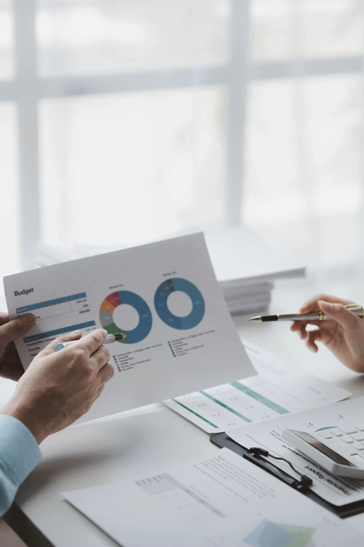 Two people review a printed budget report with charts at a desk covered with documents, a pen, and spectacles.