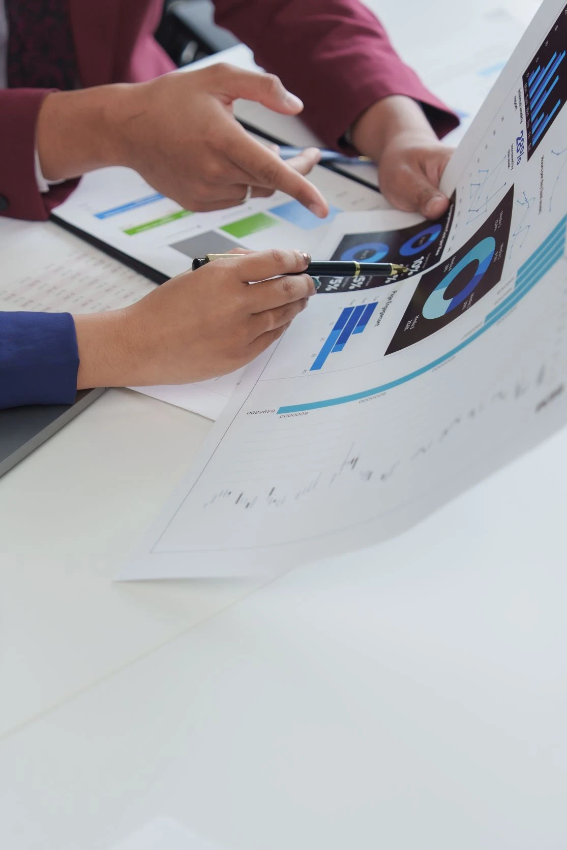 Two people review financial charts and graphs on paper, discussing data trends with pens in hand at a desk.