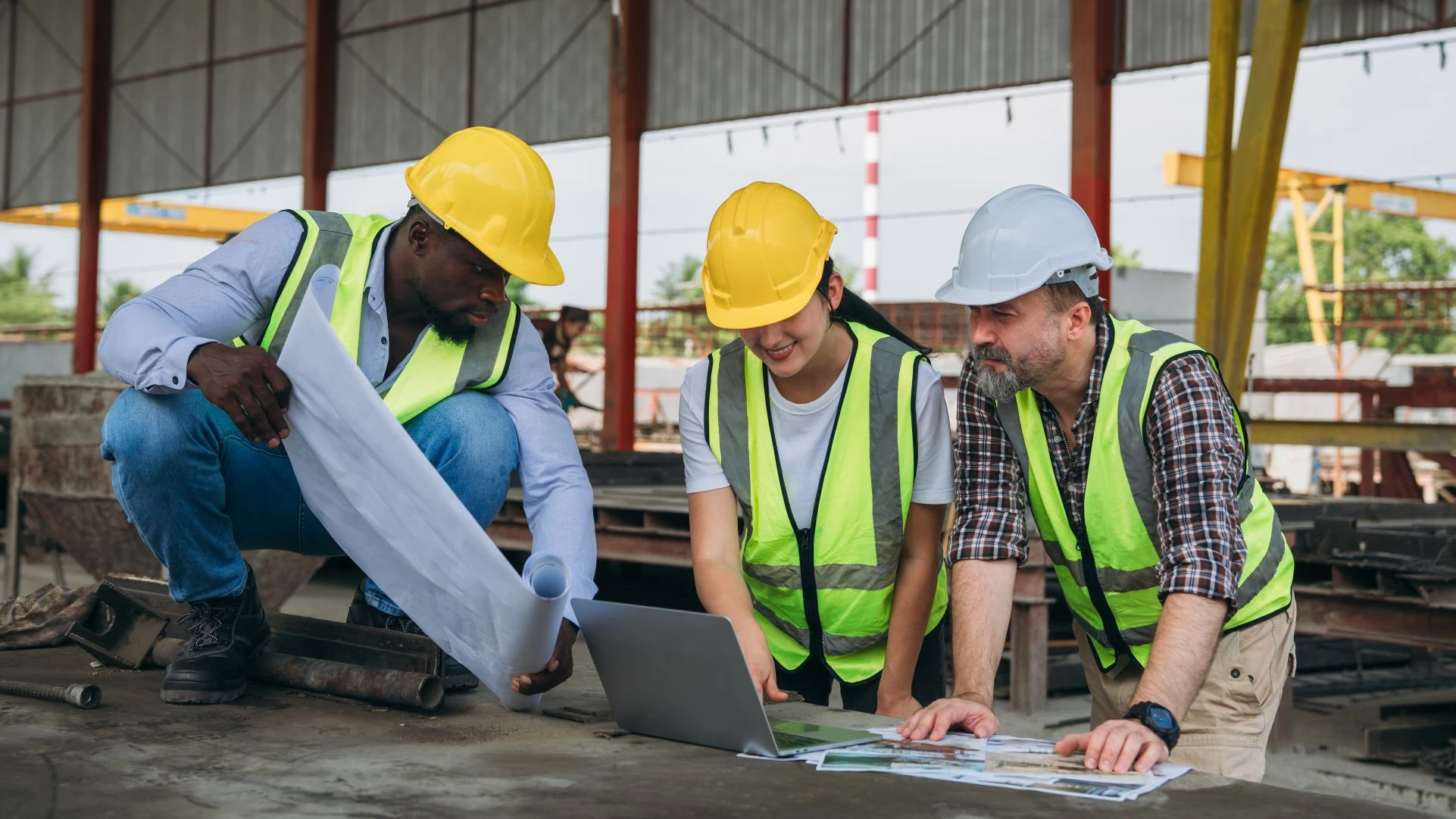 Three construction workers in safety vests and helmets review blueprints and look at a laptop on a table at a building site.