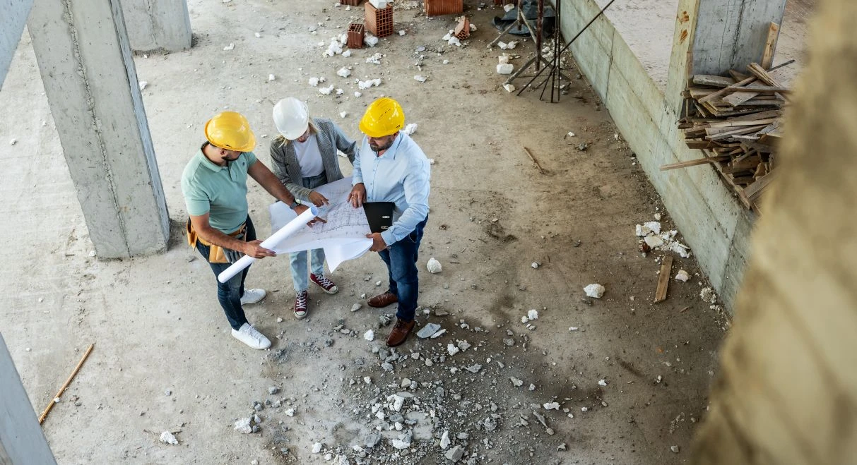 Three people wearing hard hats examine blueprints at a building site, standing on a concrete floor scattered with debris and building materials.