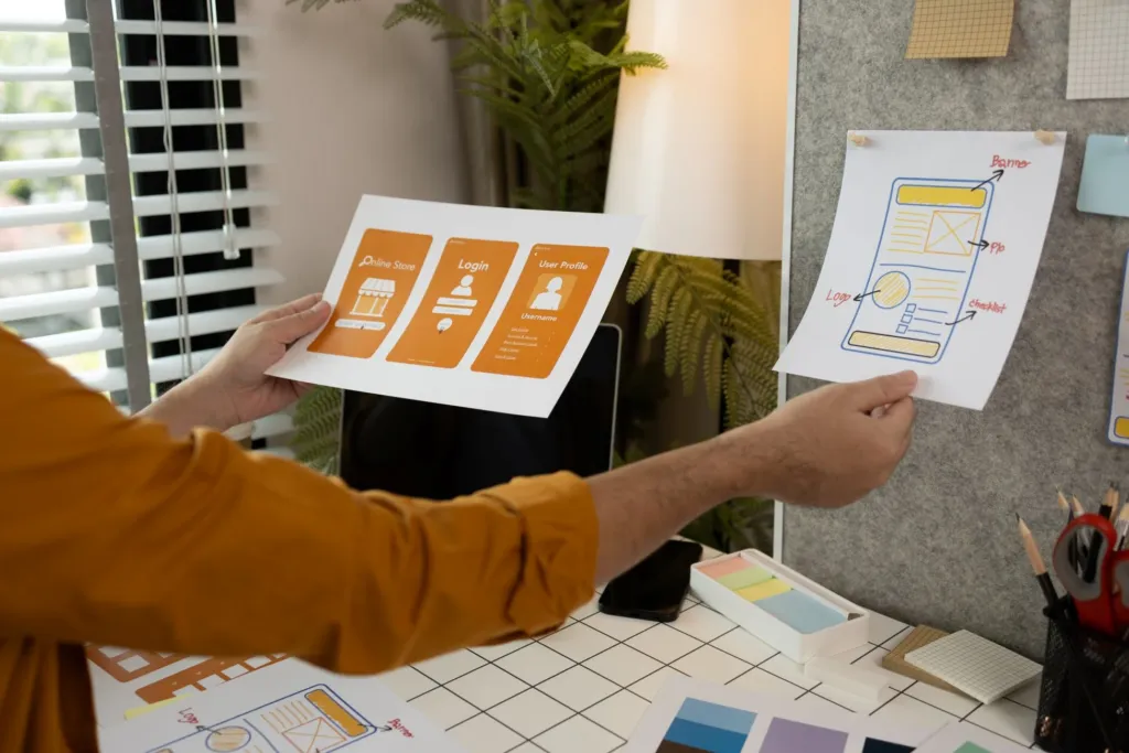 Person holding UX design sketches for a mobile app login screen and wireframe, with design materials and a computer on the desk in an office setting.
