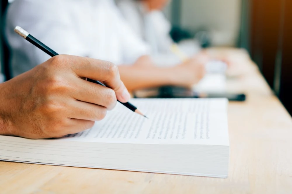 A person holds a pencil and writes in a book on a wooden desk, with another person working in the background.