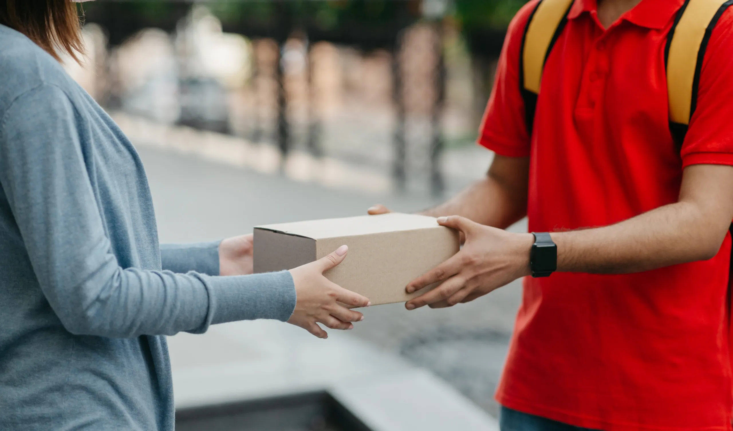 A person hands a cardboard box to another person outdoors; only their torsos and arms are visible.